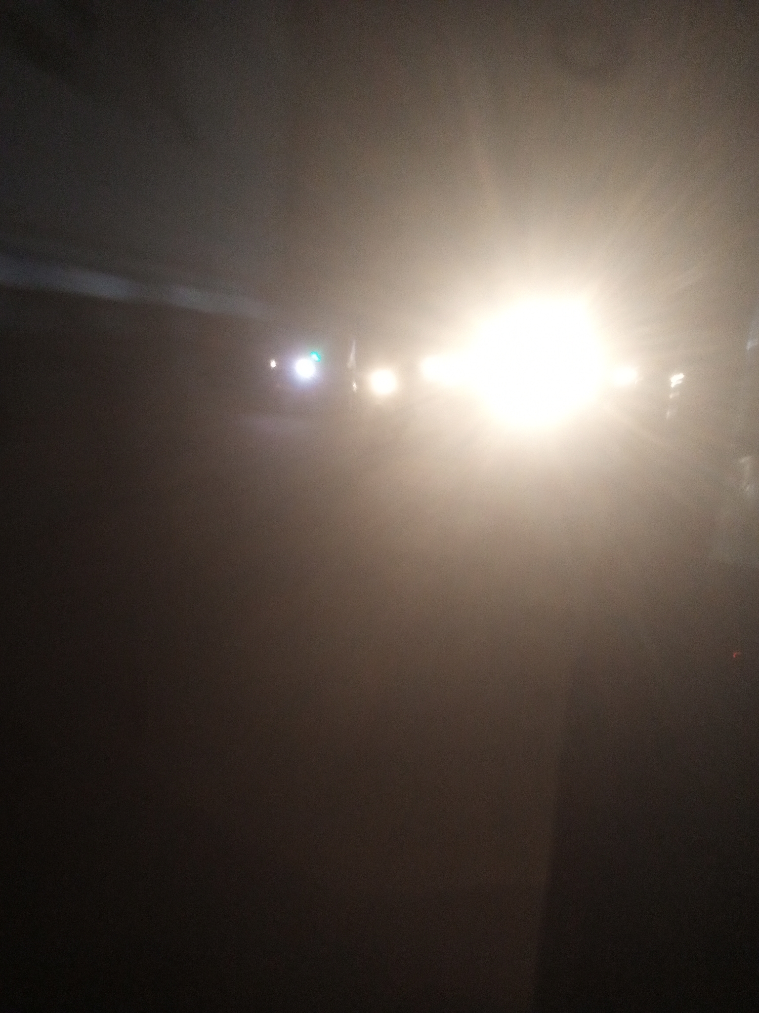 The image depicts a street scene at night, likely in Tundun Wada West, Nigeria. The dominant feature is an extremely bright, blown-out light source in the center-right of the frame, suggesting a strong headlight or streetlight. Several other, less intense lights are visible, some appearing as small white or green dots in the background, possibly indicating other vehicles or streetlights. The overall atmosphere is hazy or foggy, obscuring details of the surroundings and any potential figures. The ground appears dark and wet, reflecting some of the ambient light. The scene conveys a sense of low visibility, typical of nighttime driving in adverse conditions.