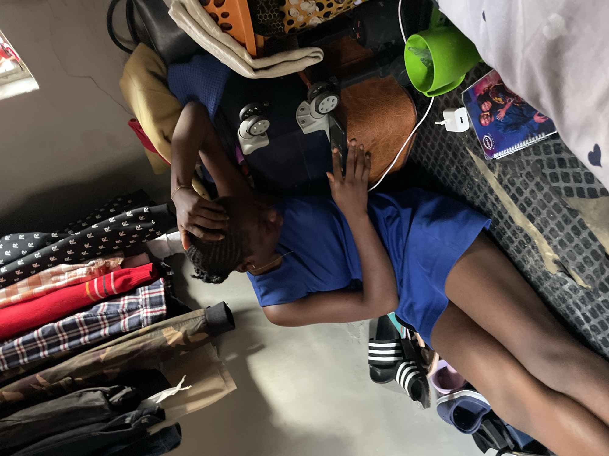A young Black woman in a blue sleeveless dress sits on a cluttered bed, looking down at her phone. She has her right arm raised, touching her hair. Several pairs of shoes are scattered around her, including a pair of black and white Adidas slides and some blue and pink slip-on shoes. Clothes are hanging on a rack to her left, and a pile of blankets, bags, and luggage is to her right. A green cup, a phone charger, and a notebook with a picture of a couple are on the bed near her. The room appears to be dimly lit with a single light source coming from a window above.