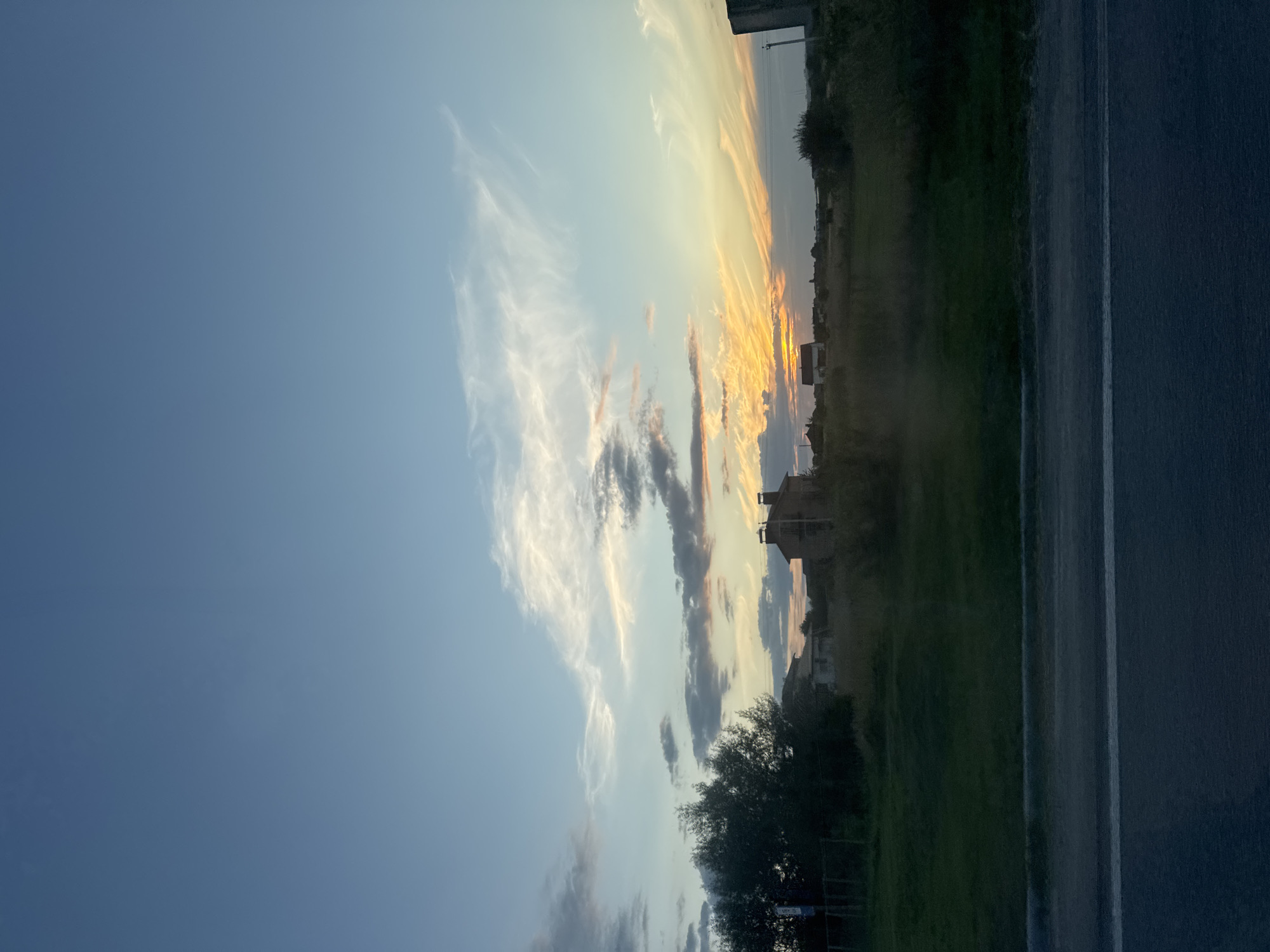 The image captures a rural sunset scene in Kopystyn, Ukraine. The sky is a blend of blues, with wispy white clouds and streaks of orange and yellow from the setting sun concentrated near the horizon. In the mid-ground, several modest houses with brick chimneys are visible amidst vegetation. A large, dark tree stands to the left, with a fence line running behind it. The foreground consists of a dark asphalt road with a white line, suggesting it is a rural road or highway. The overall impression is of a tranquil evening in a village setting.