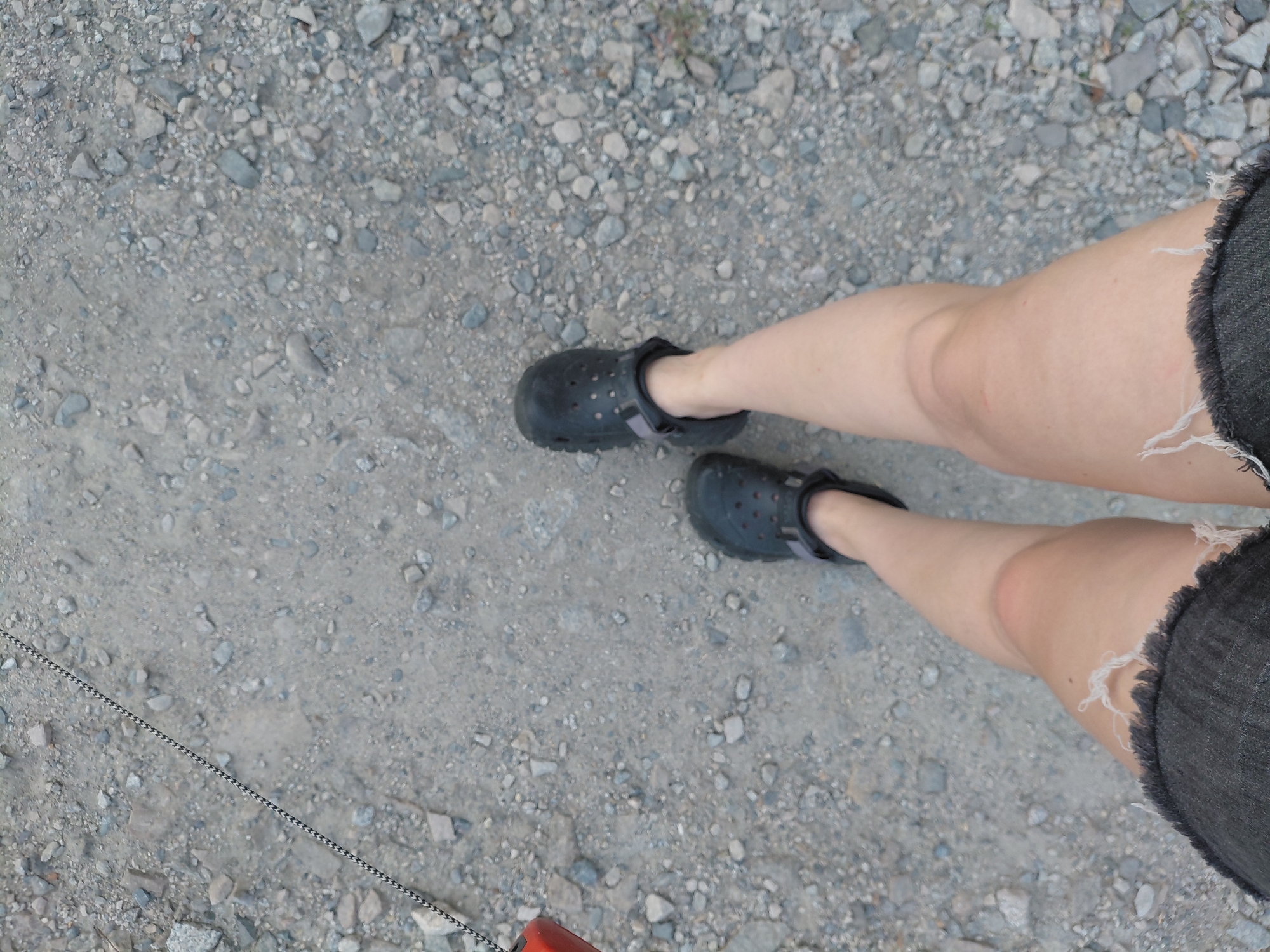 A pair of legs wearing black Crocs and distressed denim shorts are visible from a high angle, walking on a gravel path. A red object and a black and white patterned rope are partially visible on the left side of the frame. The scene appears to be outdoors, with no specific landmarks to indicate a location in Novosibirsk, Russia.
