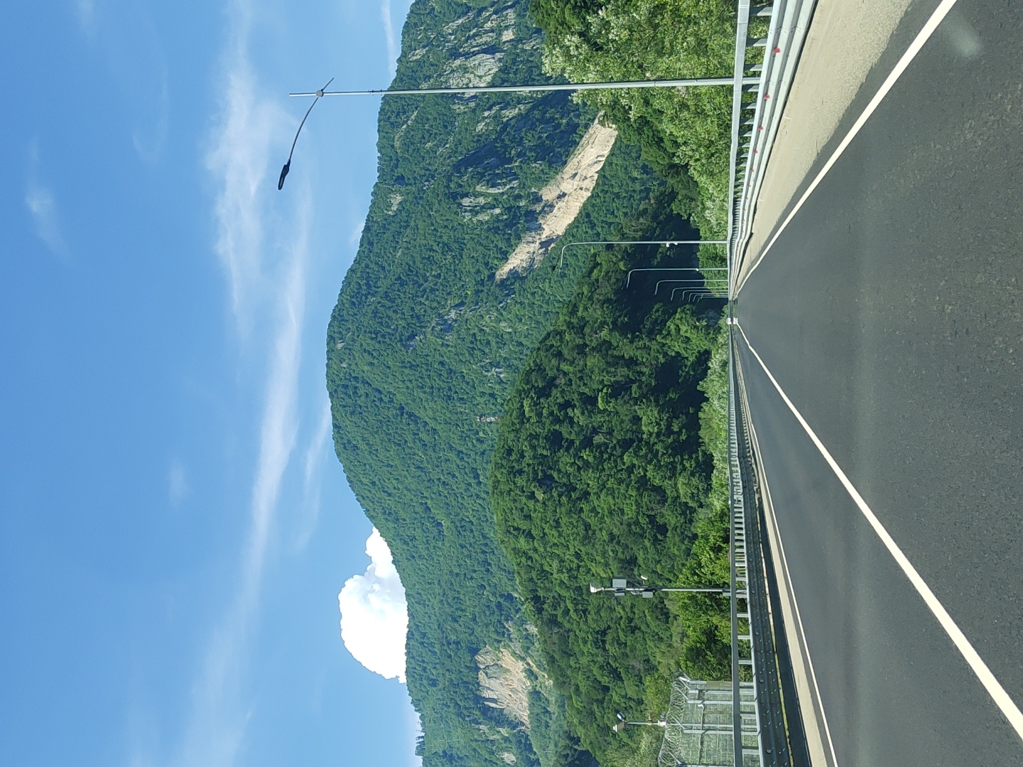 The image captures a view from a highway, likely in or near Monastyr, Russia, given the context. The road, paved in dark asphalt, is marked with white lane dividers and stretches towards a large, verdant mountain range. The mountains are densely covered with trees, with some rocky, lighter-colored sections visible on their slopes, suggesting potential geological activity or erosion. A bright blue sky with scattered white clouds forms the upper portion of the scene. Along the sides of the highway, metal guardrails are prominent. Several streetlights with dark fixtures are spaced along the roadside. To the left, a security fence with barbed wire is also visible. The overall impression is a scenic, remote highway passing through a mountainous, forested region.