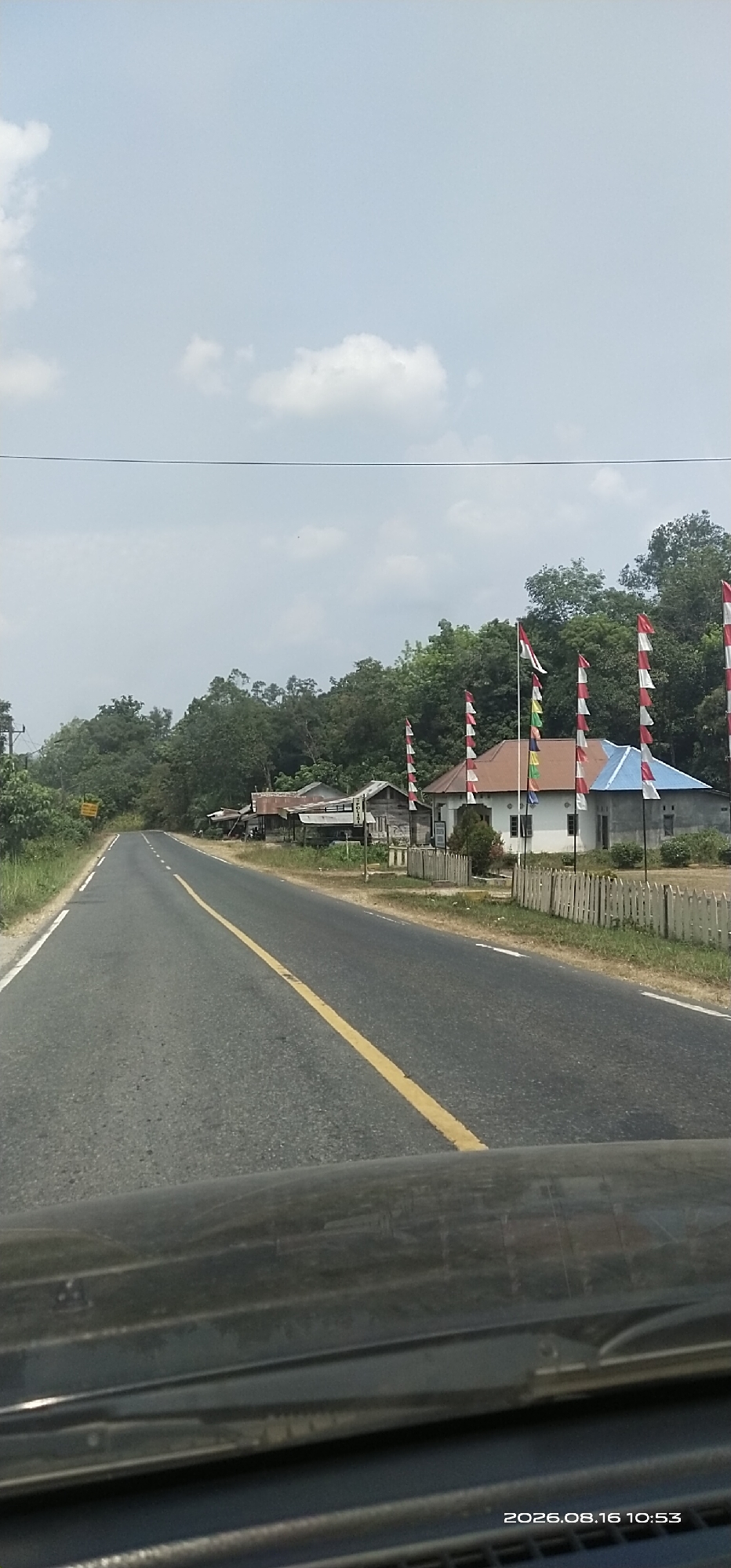 The image depicts a roadside scene with a paved road running through a rural or suburban area. The road has a yellow center line and white edge lines. On the right side of the road, there are several houses with red and blue roofs, some appearing to be simple residential structures. In front of these houses, there are rows of red and white striped flags on poles, suggesting a festive or commemorative occasion. A white picket fence runs along the edge of the property. Behind the houses, a dense line of green trees forms a backdrop. The sky is light blue with scattered white clouds. The bottom of the image shows the reflection and wiper blades on a vehicle's windshield, indicating the photo was taken from inside a car. The timestamp on the image reads "2026.08.16 10:53". The location is identified as Tampa, Indonesia.