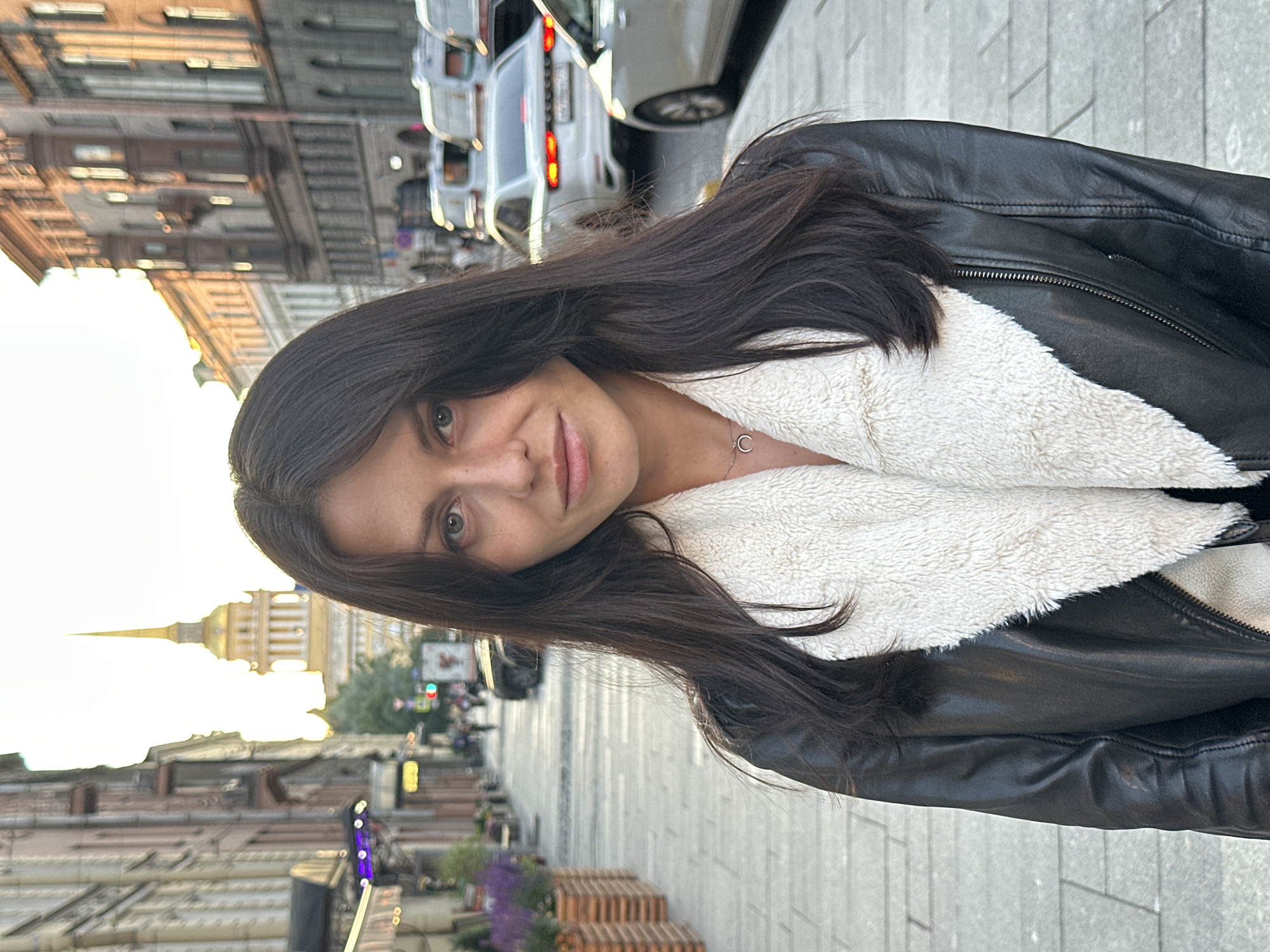 The image features a young woman with dark hair, wearing a black leather jacket lined with white fluffy material. She is looking towards the camera with a slight smile. Her face is angled, with her left eye more prominent. She is wearing a simple silver necklace with a pendant.
The background suggests an urban setting, possibly in Saint Petersburg, Russia, given the architectural style of the buildings visible and a prominent golden spire with a dome in the distance, reminiscent of the Peter and Paul Fortress or Admiralty Building. The street is paved with grey interlocking stones, and there are cars parked or driving on the right side of the frame. The lighting indicates it is either early morning or late afternoon, with the sun casting a warm glow on the buildings. Some streetlights and a traffic light are also visible. There are planters with purple flowers adding a touch of color to the scene. The overall mood appears casual and relaxed.
