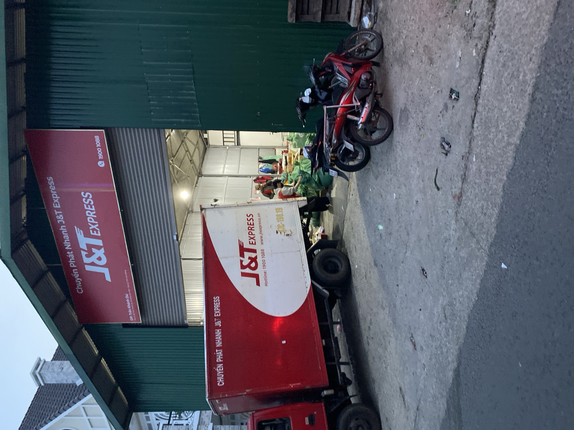 The image shows a J&T Express logistics operation, presumably in the Lâm Viên - Đà Lạt, Vietnam area. A J&T Express truck is parked, likely for loading or unloading. Two motorbikes are parked nearby. Inside the warehouse, people are visible, likely sorting or handling packages. The J&T Express signage is clearly visible on the building and the truck. The environment appears to be a typical warehouse/industrial area.