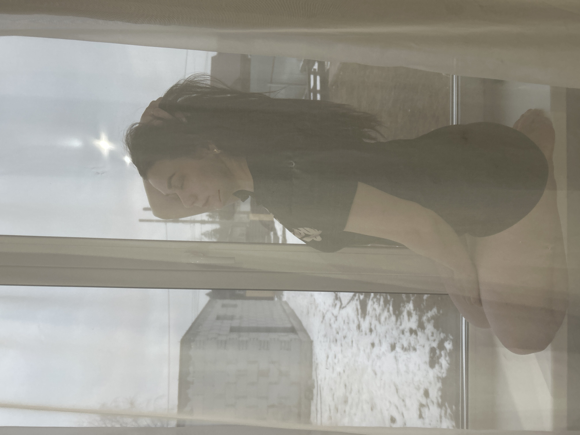 A woman with long, dark hair pulled back is kneeling on a windowsill inside a building in Chaikovsky, Russia. She is wearing a dark-colored t-shirt and shorts. Outside the window, a tall, light-colored building is visible, along with trees and sky. The photo appears to have been taken through a dirty or frosted window, obscuring the view slightly.