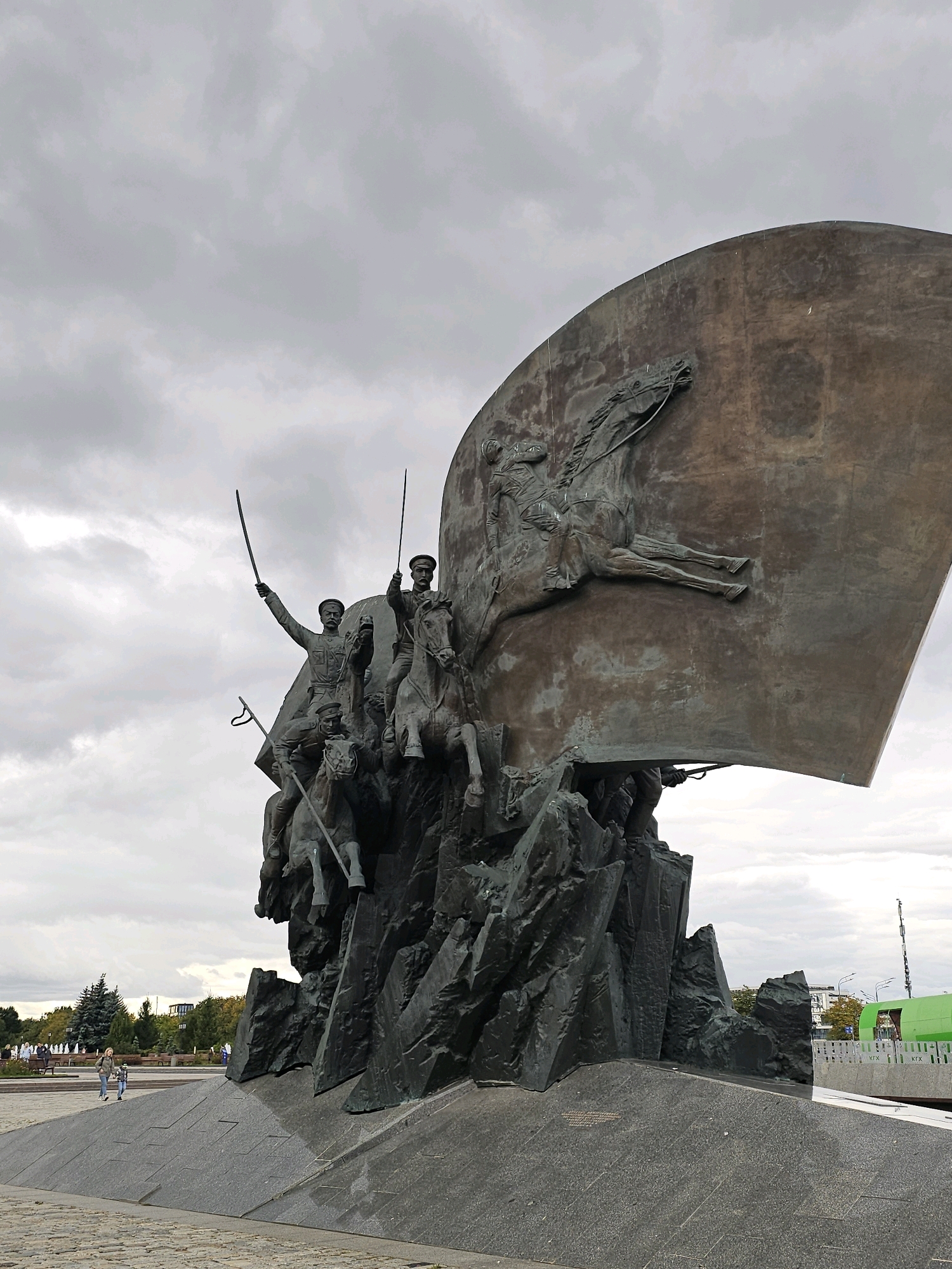 The image captures a portion of the Monument to the Leaders of the White Movement in Moscow, Russia, under an overcast sky. The monument features a large, curved metal structure adorned with bas-relief sculptures of figures on horseback. In the foreground, a group of bronze figures on horseback are prominently displayed, appearing to surge forward. These figures are depicted with swords raised. The entire sculpture is mounted on a dark, jagged rock-like base, which rests on a granite platform. To the left of the monument, two individuals, a woman and a child, are walking across a paved area. In the background, trees, benches, and a distant green structure are visible.