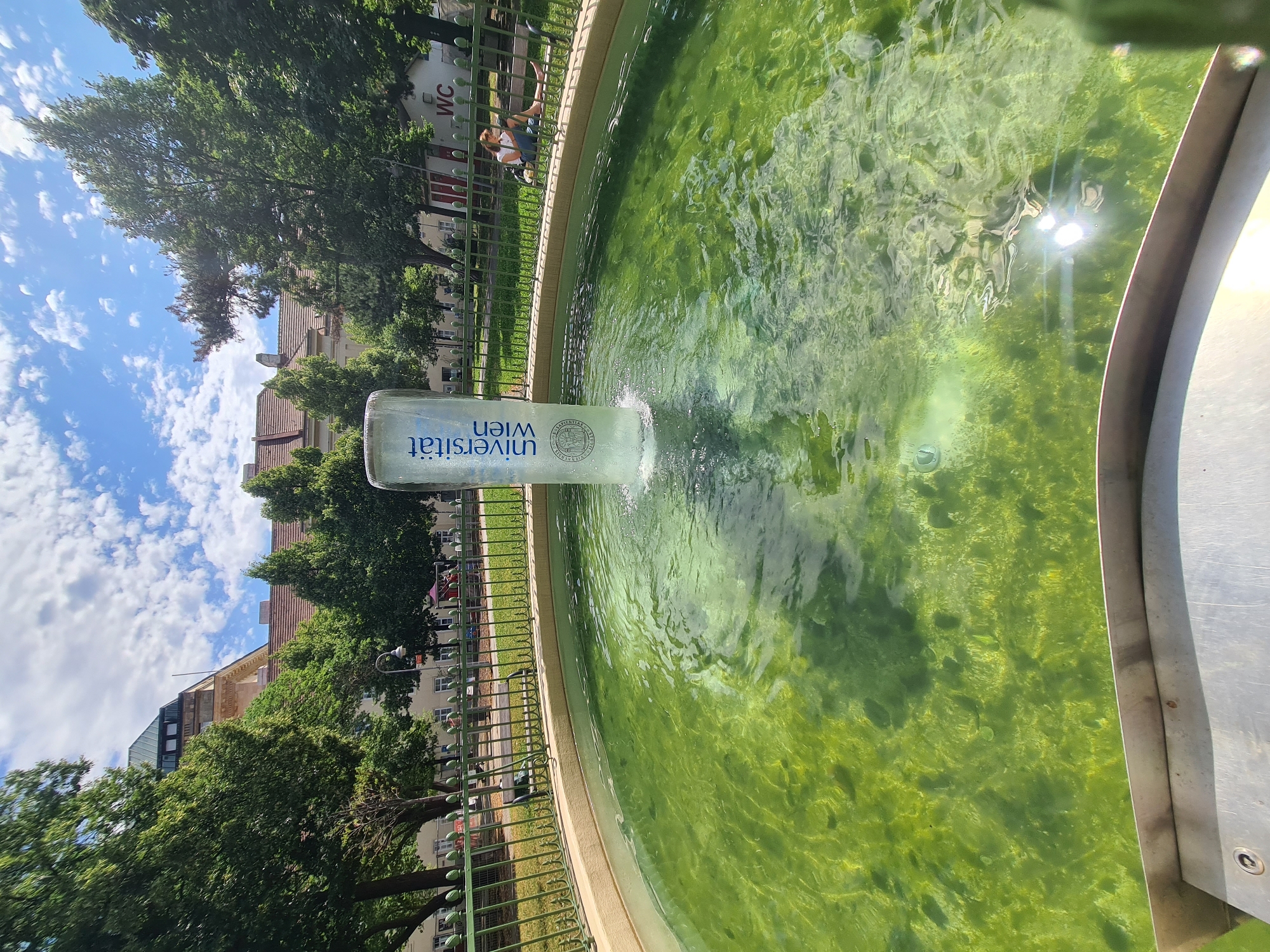 A water fountain with green, algae-filled water is the main subject of this image, taken outdoors in Vienna, Austria. The fountain is made of clear glass and has "Universität Wien" and a crest printed on it. The fountain is surrounded by a beige-colored rim, and behind it is a green metal fence.
Beyond the fence, there are trees, buildings, and a blue sky with white clouds. A person is sitting on a bench behind the fence, reading a book. There is also a building with a "WC" sign on it in the background. The scene is well-lit by the sun, which creates reflections on the water's surface. The overall impression is of a sunny day in a park or courtyard.