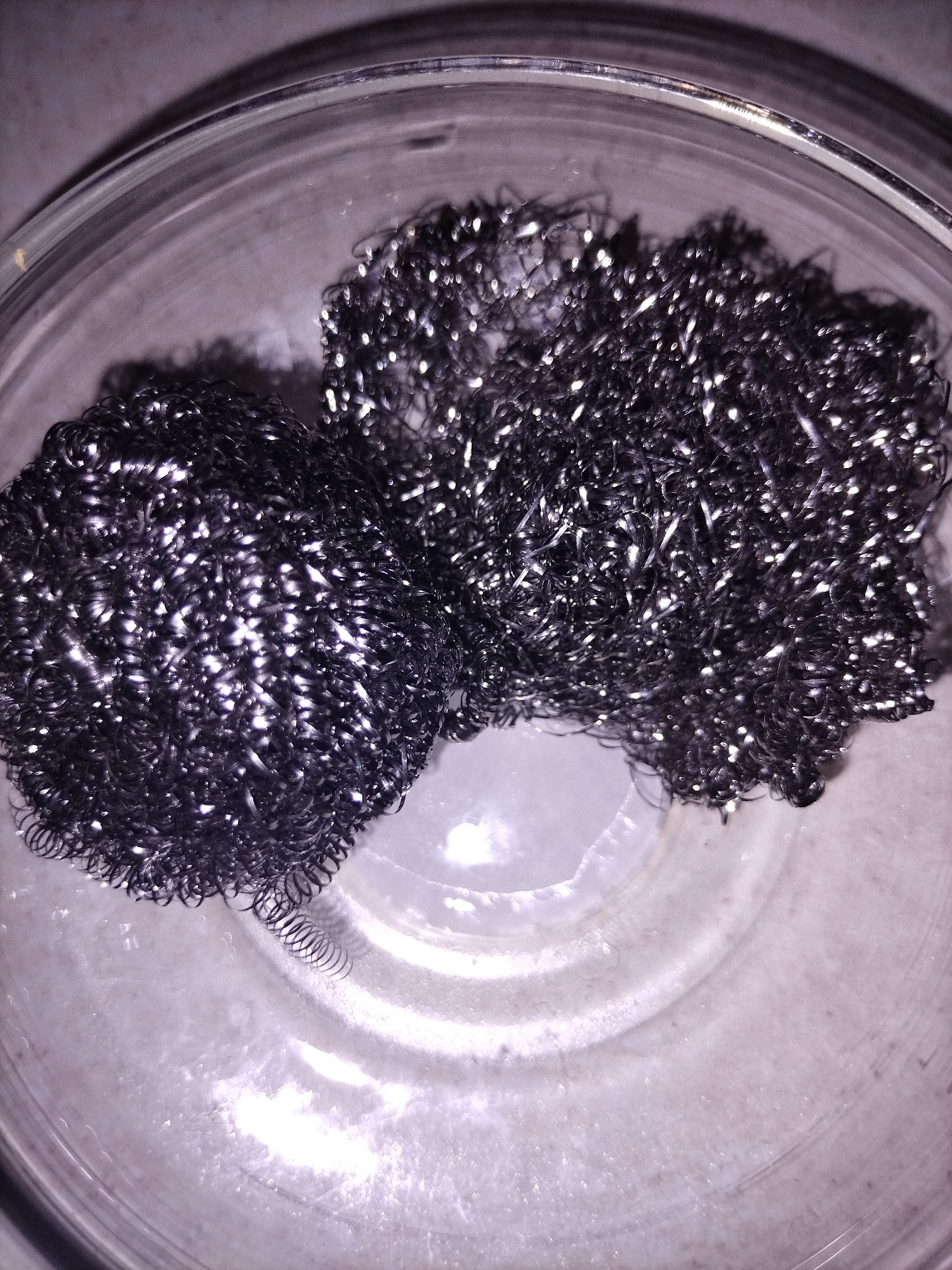 A close-up, overhead shot depicts two steel wool scouring pads resting inside a clear glass bowl. The scouring pads are dark gray and have a rough, metallic texture, with light reflecting off their surfaces. The glass bowl is transparent, revealing a light-colored, textured surface underneath, which appears to be a countertop. The lighting in the image is bright, creating highlights and shadows that emphasize the texture of the scouring pads and the bowl. The scene is static and there are no people or animals present. The location is unknown.
