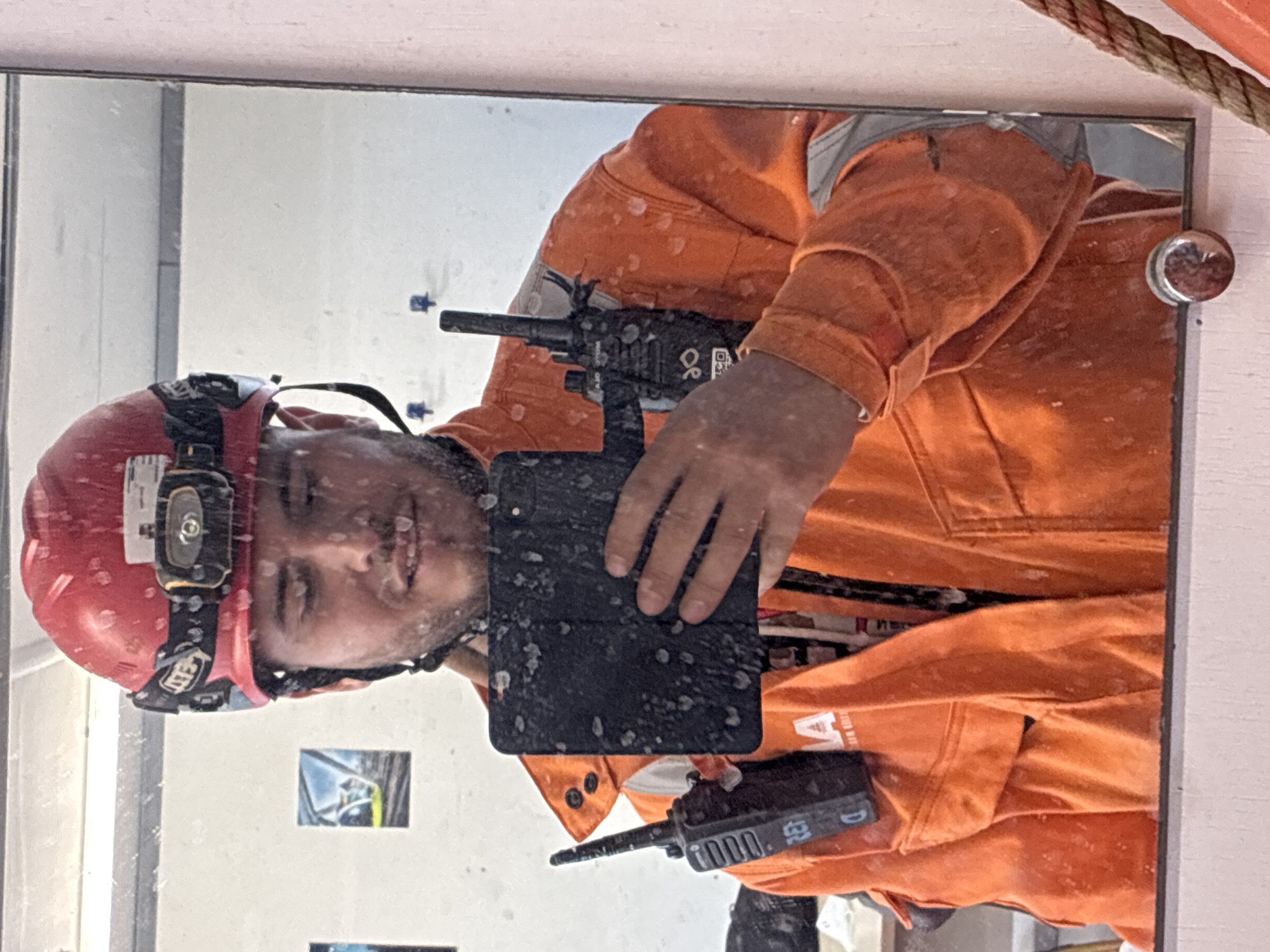 The image depicts a man wearing an orange uniform and a red helmet with a headlamp, taking a selfie in a mirror. The mirror appears to be mounted on a white door or wall. The man is holding a black tablet in front of him, and a handheld radio is visible both above and below the tablet. The man is likely a worker, possibly in construction or a similar field. The image shows white specks all over the man and mirror. The location in the city of Brest, France is not discernible from the image.