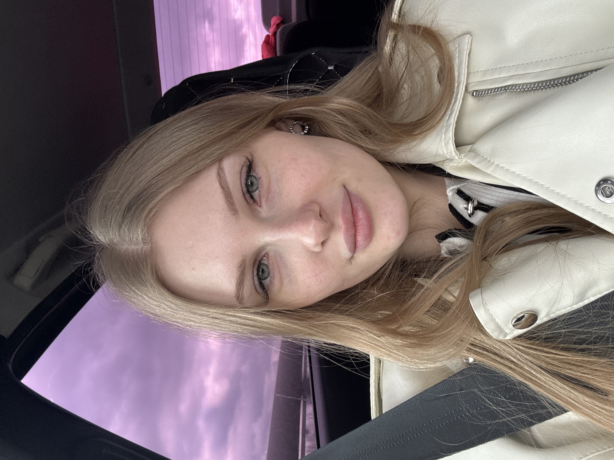 A young woman with blonde hair and fair skin is in a car. She's wearing a white leather jacket and a black and white striped shirt. A black seatbelt is visible across her chest. Outside the car window, a purple sky can be seen, indicative of dawn or dusk. The location is not discernible as Perm, Russia from the available visual information.