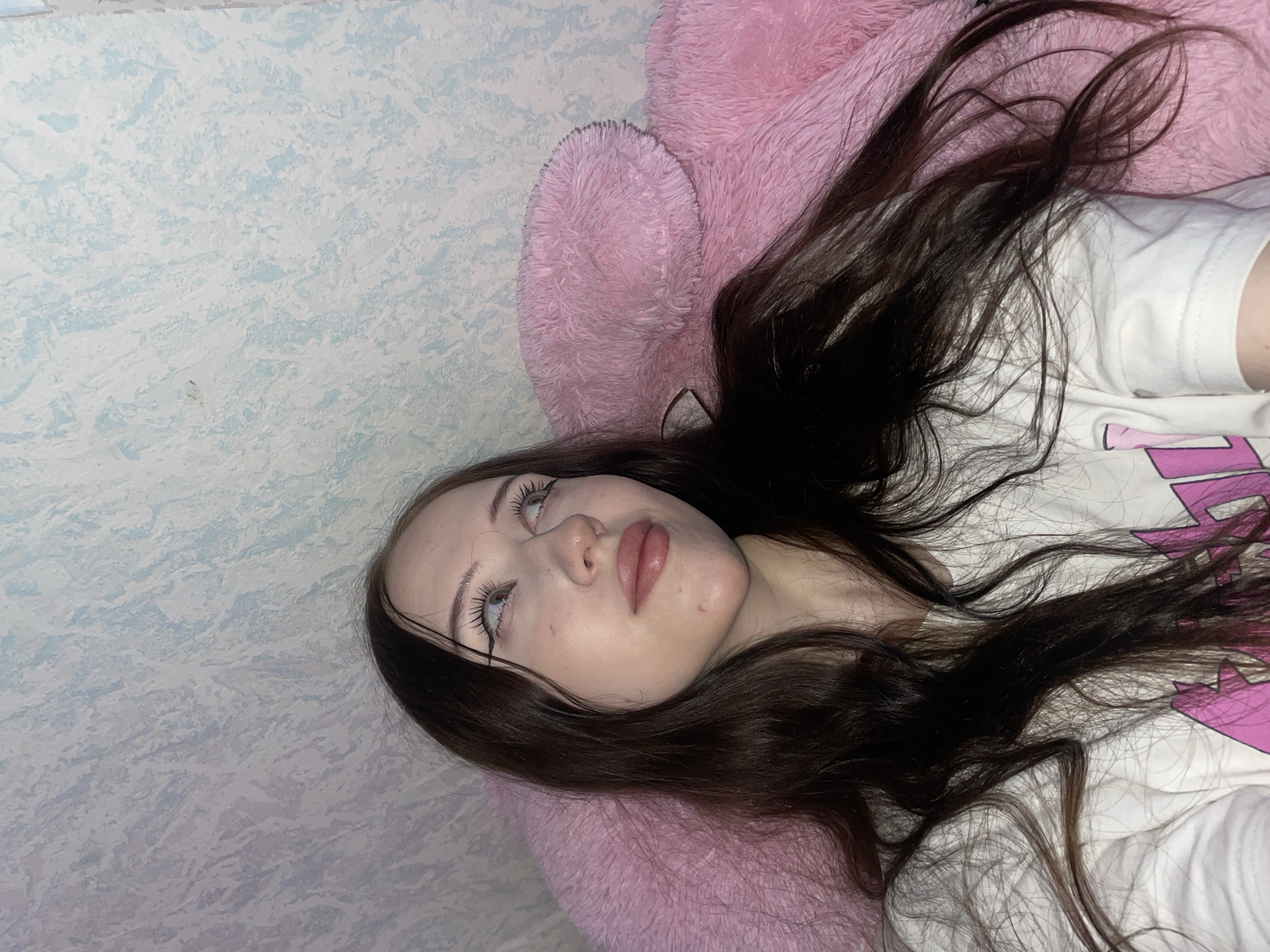 The image shows a young woman with long, dark brown hair, looking upwards and to the right. She is wearing a white t-shirt with pink lettering. She has noticeable makeup, including winged eyeliner and lipstick. She is lying on a large, fluffy pink object, possibly a pillow or stuffed toy, against a textured blue and white wall. The scene appears to be indoors. There is no visible indication of the city of Votkinsk, Russia in the image.
