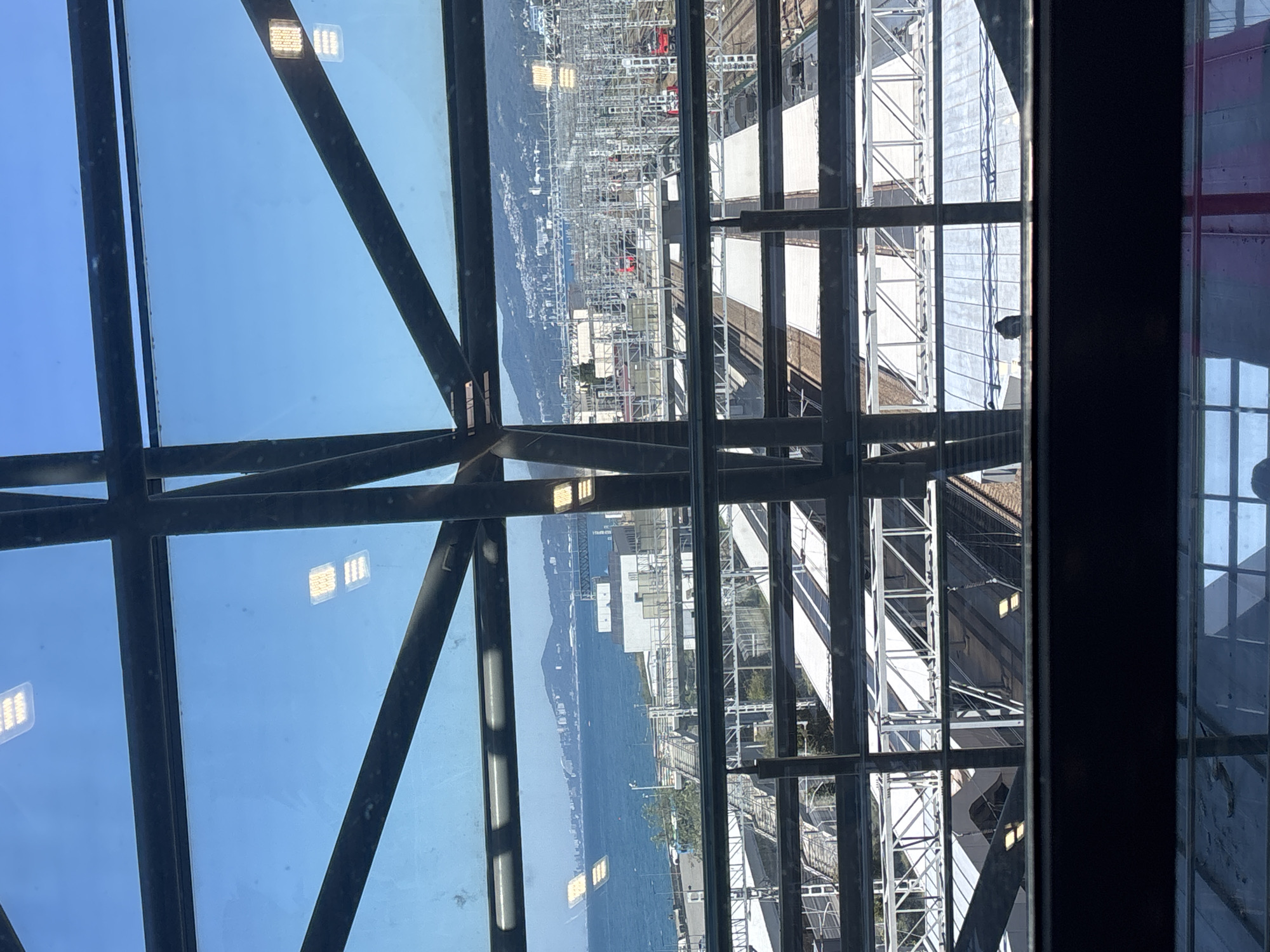 The image provides a view from inside a structure with large glass windows and dark metal framing. Through the windows, a cityscape is visible, dominated by a large body of water, likely the Black Sea, and a distant shoreline with buildings and mountains. In the foreground, there are multiple train tracks and platforms, indicating a railway station. The sky is clear and blue. Some lights are visible on the ceiling of the interior structure. The overall scene suggests a modern transportation hub situated near the coast in Sochi, Russia, possibly at the Sochi Railway Station or a related facility offering elevated views of the area.