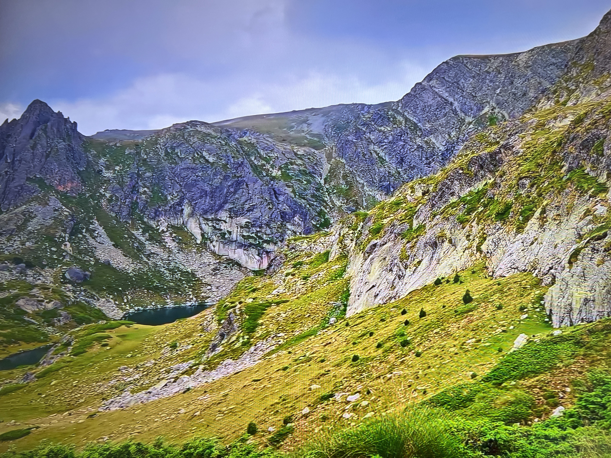 The image shows a mountainous landscape with two small lakes in the lower left quadrant. The mountains are a mix of rocky terrain and grassy slopes. The sky is partly cloudy. The location is in the city of Unknown, Unknown.