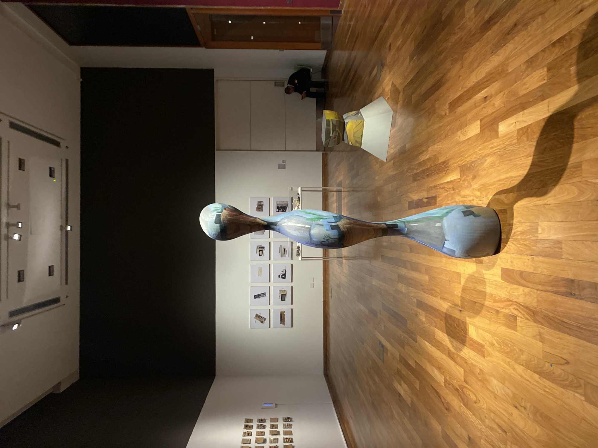 The image depicts an art gallery exhibit, likely located indoors in a building with hardwood floors. A large, hourglass-shaped sculpture with a blue and brown design dominates the foreground. On the wall behind the sculpture, a series of framed photographs are displayed. Further back, in the upper left of the frame, are more photographs on a wall and the exhibit has a solid black wall. In the far corner, a person is sitting. The room appears to be well-lit, casting shadows from the sculpture. The specific location is an art gallery, but the city of Buenos Aires, Argentina cannot be confirmed from the provided image.