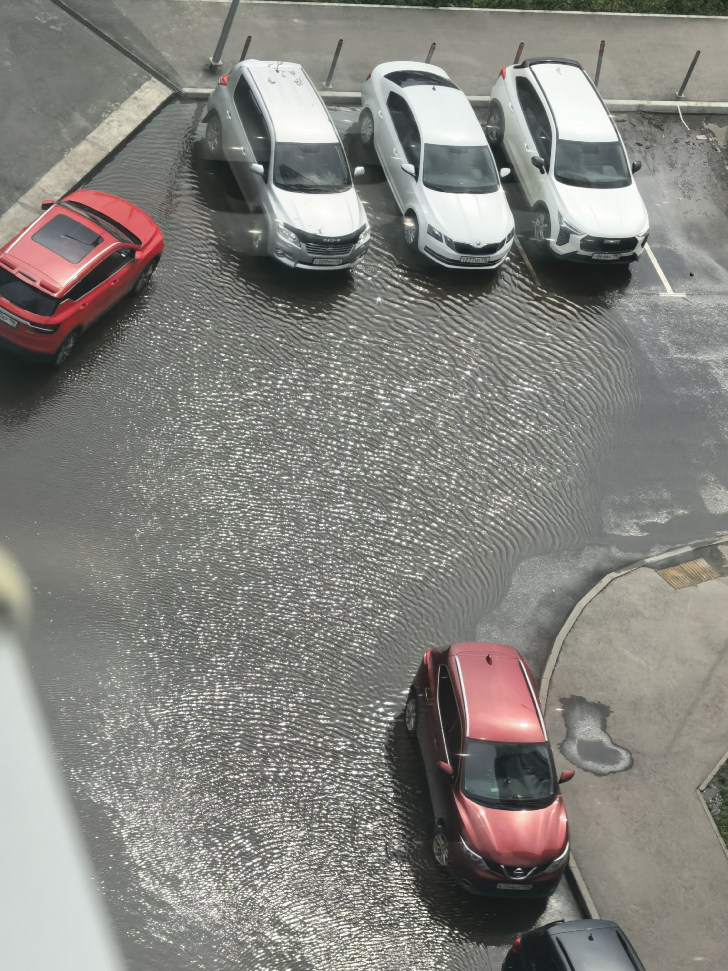 An overhead, high-angle, medium shot captures a flooded parking lot with several cars partially submerged in water. The water creates ripples and reflects the sunlight, indicating it is likely daytime.
In the upper left quadrant of the image, a red SUV with a sunroof is parked. To its right, a silver SUV is parked in the water. Further to the right, two white sedans are also in the flooded area, one slightly behind the other. The front sedan has its tires submerged, and the rear sedan is also partially inundated.
In the lower right quadrant, a red hatchback is parked on the edge of the flooded area, with its rear tires in shallow water. A black vehicle is parked behind it, with its front tires visible on dry pavement.
The parking lot appears to be paved with asphalt, and a curb separates the flooded area from a sidewalk or pathway. There are also some posts or bollards visible in the flooded section. The overall scene suggests a significant amount of rainfall has caused localized flooding.