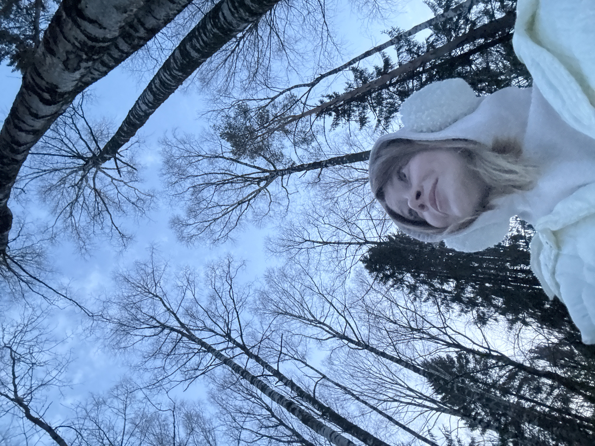 The image is a low-angle selfie taken outdoors in Вассята, Russia. A person with fair skin, light blonde hair, and wearing a white hooded jacket with pom-poms is looking down at the camera. The background consists of a dense forest of tall, leafless trees, predominantly birch with visible black and white bark patterns. Some evergreen trees are also present. The sky is visible through the trees, appearing light blue and slightly overcast. The scene suggests a winter setting in a wooded area.