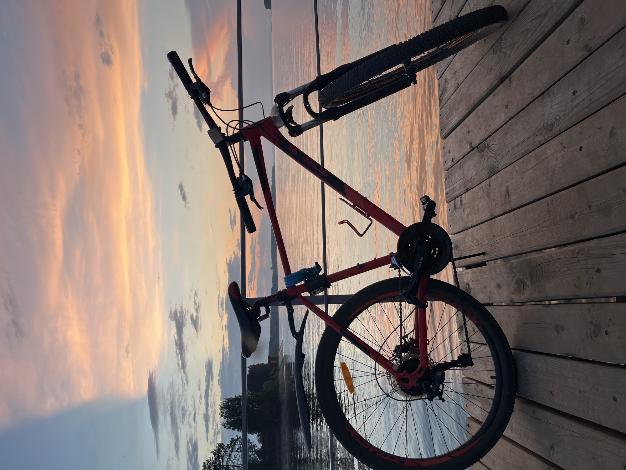 A red bicycle is parked on a wooden pier, facing right. The bicycle has a blue lock on its frame and a water bottle cage. The pier is situated beside a calm body of water, reflecting the colors of a sunset. The sky above is a mix of blue, pink, and orange hues with scattered clouds. Trees are visible in the background on the opposite shore. This scene is located on the Kama River embankment in Chaikovsky, Russia. There are no people or animals present in the image.