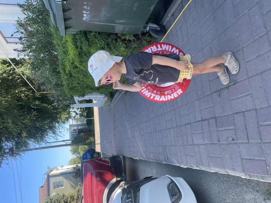A young child stands in profile on a paved brick sidewalk in Starotitarovskaya, Russia, facing right. The child wears a light-colored bucket hat with a floral design, pink oval sunglasses, a dark t-shirt with a light graphic, yellow and white checkered ruffled shorts, and light-colored closed-toe sandals. A red inflatable swimming ring, labeled "SWIM TRAINER" in white text with additional safety markings, is positioned around the child's waist. The child's right hand is raised to their chin.
To the left of the sidewalk, a white vehicle's front quarter is visible, followed by the rear of a red pickup truck, both parked on an unpaved or asphalt surface. A blue vehicle is partially visible further down the road/driveway behind the pickup. On the right side of the sidewalk, a large, dark green wheeled waste bin stands behind a dense green hedge. In the background, a light-colored multi-story building with a pitched roof and windows is visible on the far left. A utility pole stands upright beyond the vehicles. Further along the right side of the scene, behind the hedge, are additional trees and the top of a white ornamental fence or railing. The sky above is clear and blue, indicating daylight.