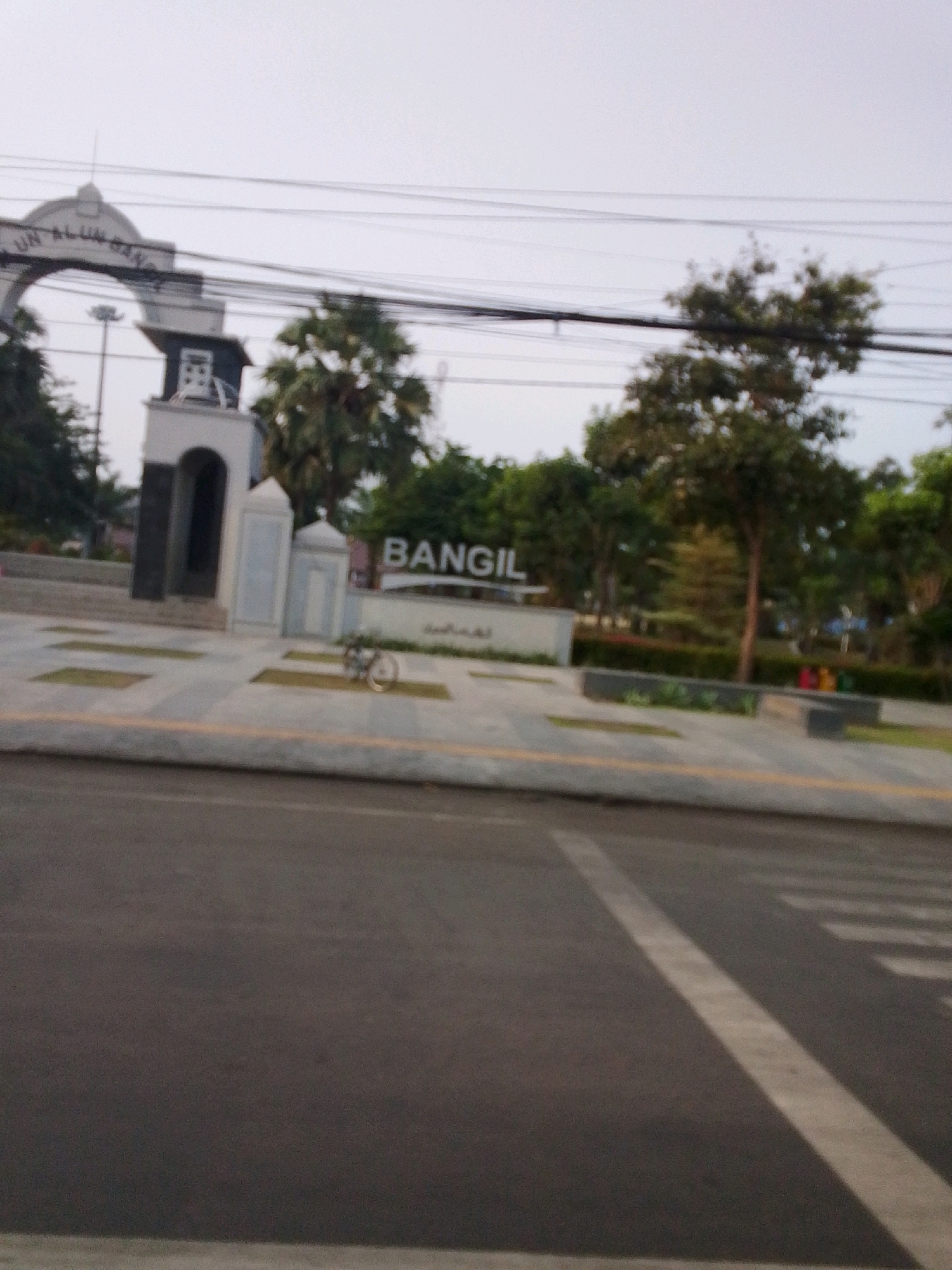 An eye-level, slightly blurred, long shot captures a scene in Bangil, Indonesia, likely near an entrance to an alun-alun (town square). In the background, a white archway with dark lettering, partially visible, stands against a pale sky. To the right of the arch is a white tower-like structure with dark accents and an arched opening. Further to the right, large white letters spell out "BANGIL". A single bicycle rests near the center of the paved area. Behind the paved area, trees and green foliage form a natural backdrop. The foreground features a grey asphalt road with white lane markings and a pedestrian crossing. Overhead, a dense network of electrical wires crisscrosses the sky. The lighting suggests an overcast day.