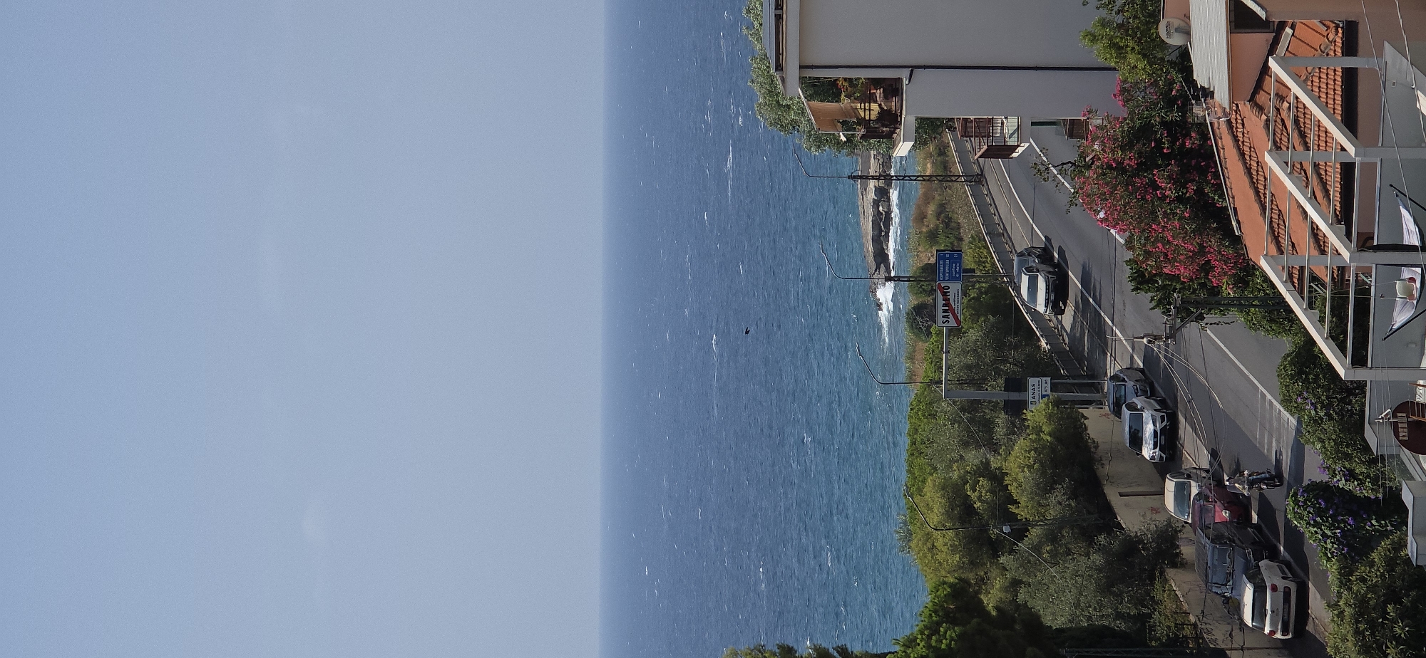 The image captures a view of the Ligurian Sea from a hillside overlooking Sanremo, Italy. The sky is a clear, pale blue. The sea is a deep blue, with whitecaps indicating a breezy condition. A small dark bird is visible in flight over the water.
In the foreground, a paved road curves uphill. Several vehicles are present: a white SUV and a darker SUV are driving on the road, while parked on the side of the road are a white hatchback, a silver sedan, a dark sedan, and a red car. A person on a scooter is also visible on the road.
A white building with multiple balconies is situated on the right side of the road, overlooking the sea. Lush green trees and bushes with vibrant pink flowers are abundant on the hillside. There are streetlights and road signs, including one indicating "SANREMO" with directional arrows to various towns and distances. A sign for "ANAS" (Azienda Nazionale Autonoma delle Strade) is also visible.
In the lower foreground, a partially visible rooftop with terracotta tiles is present, along with a white structure with a canvas hammock. The overall scene depicts a coastal urban environment in Sanremo, Italy, on a sunny day.