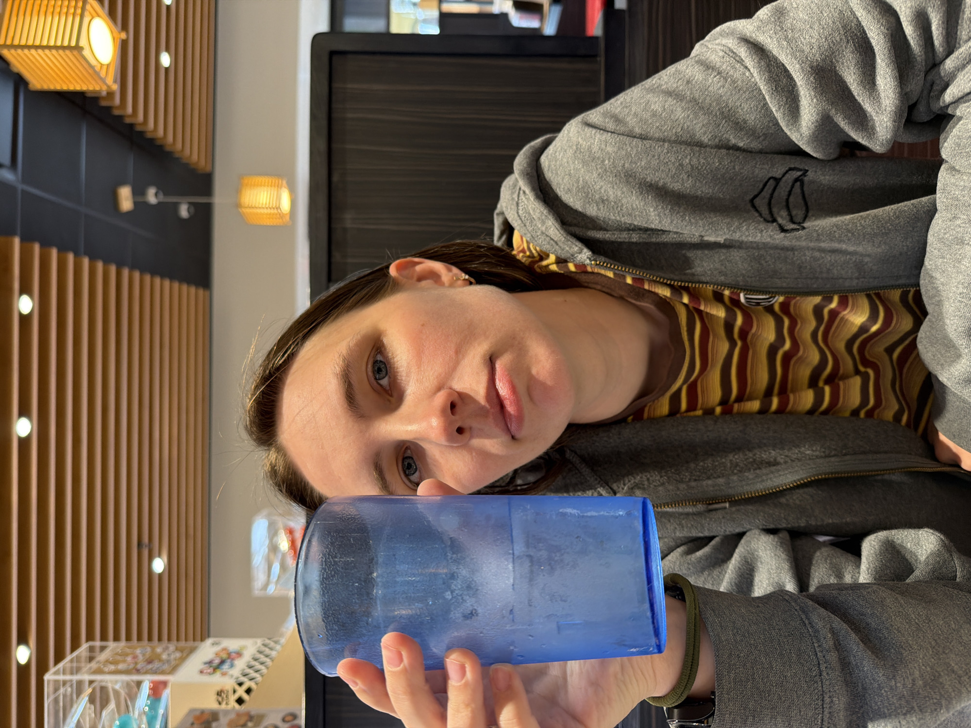 A young Caucasian woman with brown hair and blue eyes is holding up a blue plastic cup filled with ice and a clear liquid. She is wearing a grey hooded sweatshirt over a striped t-shirt with yellow, brown, and red stripes. The woman is seated at a table with a dark wooden background and a slatted wooden ceiling with modern light fixtures. The environment appears to be an indoor dining area, possibly a restaurant, with a bright, neutral color palette. This image was taken in Beaverton, Oregon, United States of America.