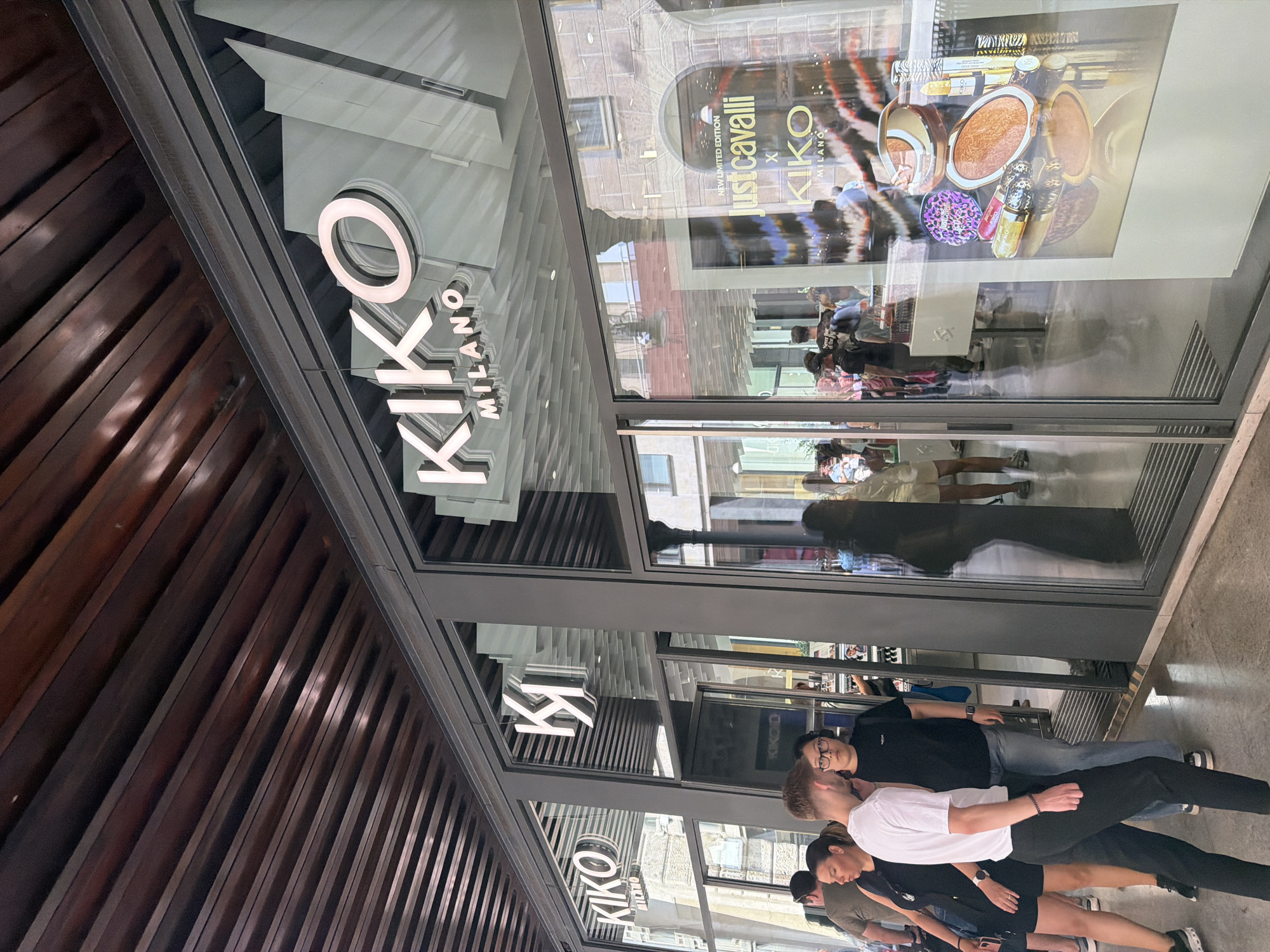 This image captures the exterior of a KIKO Milano store, a cosmetics retailer, situated in Milan, Italy. The store features large glass windows displaying various makeup products, including bronzers and lipsticks. Prominently displayed on the storefront is the KIKO Milano logo. In the foreground, several individuals are seen walking by or standing outside the store, suggesting a bustling urban environment. The architecture visible in the reflection of the windows indicates a classic European city setting. The overall scene is one of everyday street life in Milan, with a focus on a retail establishment.