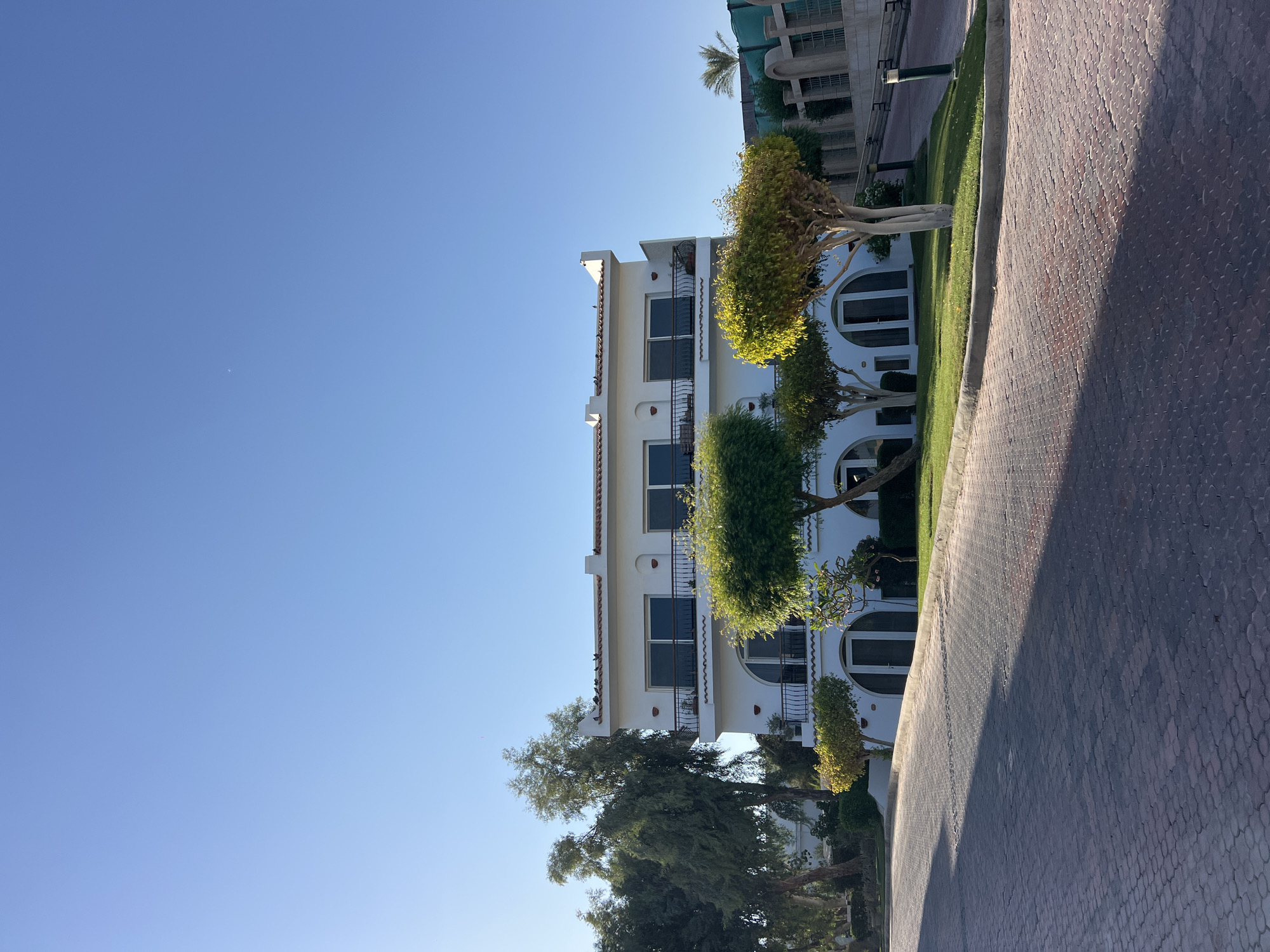 This is a full, eye-level shot of a white, multi-story building in Sharm El Sheikh, Egypt, bathed in bright sunlight. The building has balconies with black railings on both the second and third floors, and arched doorways on the ground level. Several trees with manicured canopies of green and yellow foliage are situated in front of the building. To the left of the building, taller, more natural-looking trees are visible. The foreground is dominated by a paved pathway made of interlocking red bricks, casting a long shadow. In the background, a green fence and some structures can be seen. The sky above is a clear, bright blue with a sliver of the moon visible. There are no people in the frame, and the scene appears to be peaceful and quiet.
