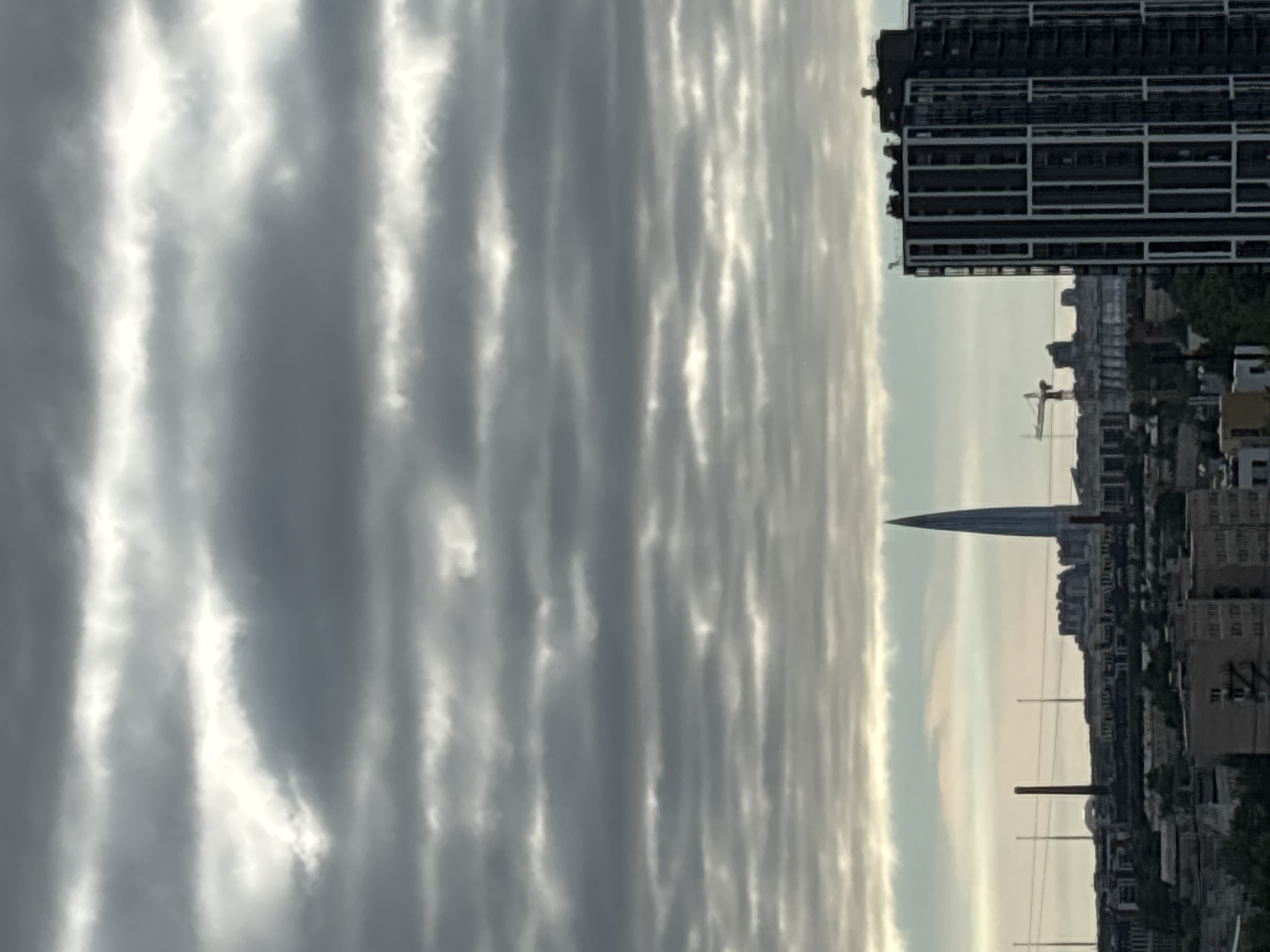 The image captures a cityscape under a cloudy sky. In the foreground, a modern, dark-colored skyscraper with white vertical accents dominates the right side of the frame. Further back, a collection of buildings forms a dense urban area. A distinctive, slender, pointed skyscraper, likely the Lakhta Center, rises prominently in the middle ground, its modern architecture contrasting with the surrounding industrial-looking structures and smokestacks. A construction crane is visible near the Lakhta Center, suggesting ongoing development. The sky is filled with horizontal layers of gray clouds, with some brighter patches indicating diffused sunlight. The overall atmosphere is somber and overcast, typical of a cloudy day in a large city.