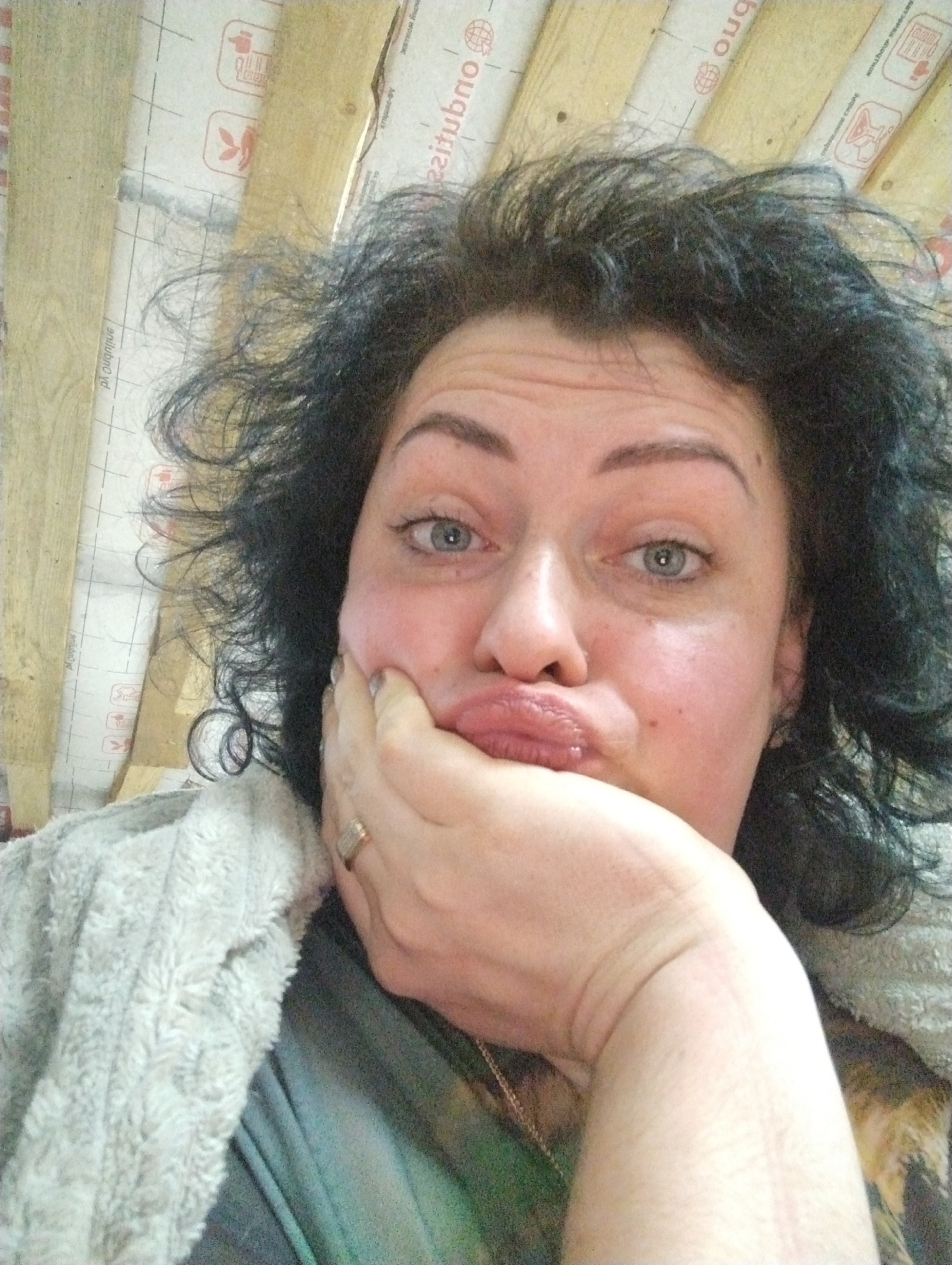 A close-up selfie of a woman with dark, curly hair and light eyes, partially obscured by her hand, is set against a background of wooden planks covered with a white, printed material, likely insulation. She is wearing a fluffy, light-colored garment, and a hint of clothing is visible underneath. The location in the city of Grishanki, Russia is not evident from the provided image.