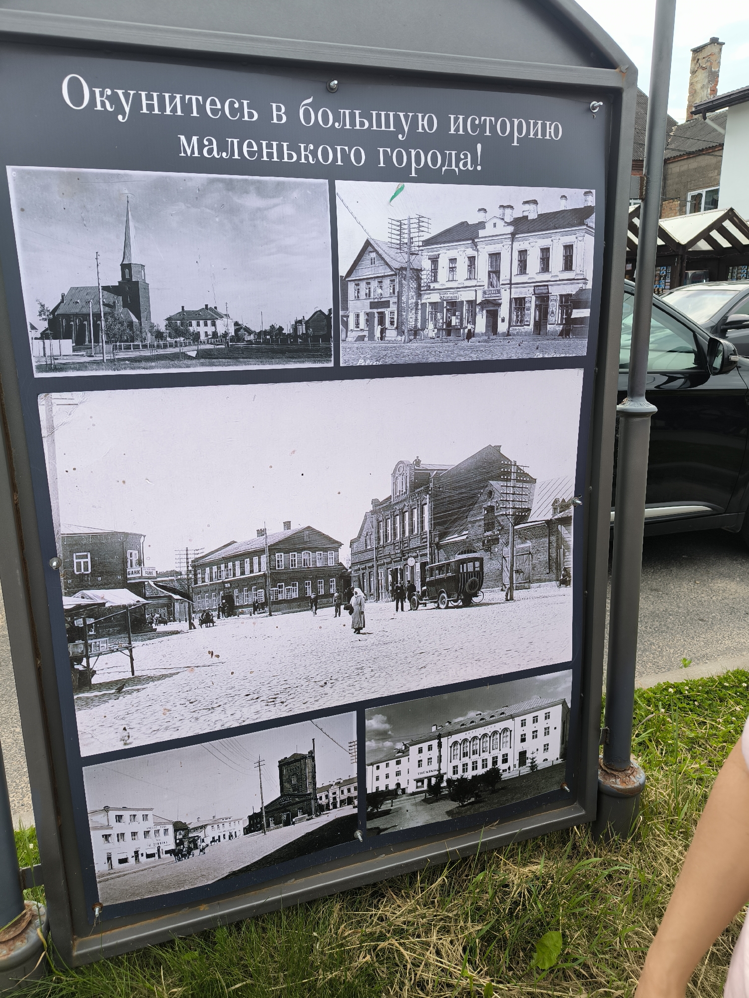 The image shows a sign with several black and white historical photographs of the city of Pechory, Russia. The sign has text in Russian that translates to "Immerse yourself in the great history of a small town!". The photographs depict various street scenes, buildings, and people from different eras, showcasing the historical architecture and daily life of Pechory. The sign is outdoors, with grass and a street visible in the background, along with a modern car.