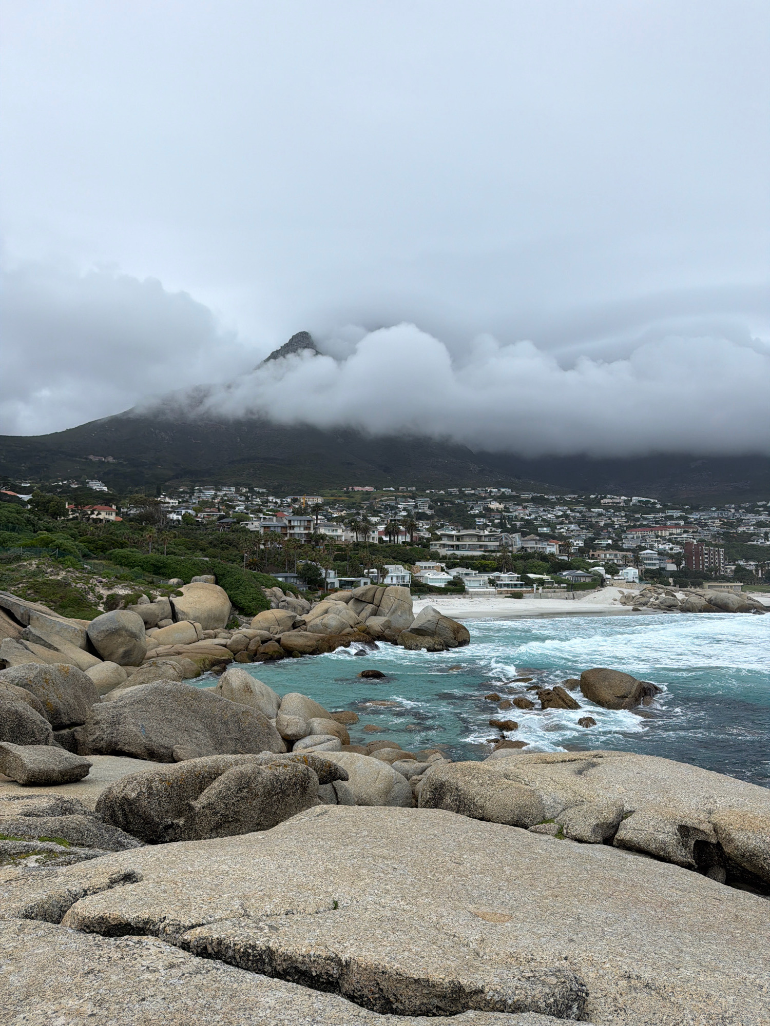 The image depicts a coastal scene in Cape Town, South Africa, likely Camps Bay or Clifton due to the presence of large granite boulders and a sandy beach backed by a hillside dotted with white and light-colored houses. The foreground is dominated by a cluster of these large, rounded granite boulders, some of which are wet and dark, indicating proximity to the water. The sea is a deep turquoise with white-capped waves crashing against the rocks and the shore.
In the midground, a white sandy beach is visible, with more of the characteristic boulders scattered along its edge. Beyond the beach, the hillside rises steeply, covered in dense green vegetation and featuring a dense collection of residential buildings. The architecture appears to be modern and Mediterranean-inspired, with white or light-colored facades and flat or tiled roofs.
The background is dominated by a large mountain, its peak shrouded in heavy, grey clouds. The clouds are low-hanging and obscure much of the mountain, creating a dramatic and somewhat overcast atmosphere. The sky itself is a uniform light grey. There are no people or animals visible in the scene. The overall impression is one of a rugged and beautiful coastline meeting a developed urban area, under a moody, cloudy sky.