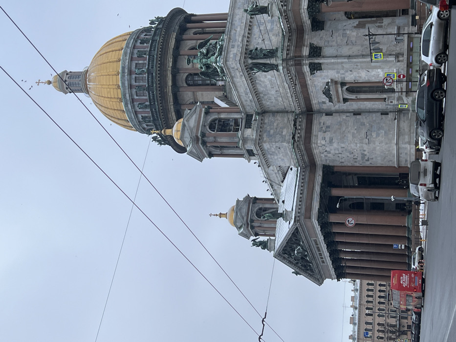 The image displays Saint Isaac's Cathedral in Saint Petersburg, Russia, under an overcast sky with light snow visible on architectural ledges and parts of the street. The cathedral is a massive neoclassical structure, centrally dominated by a large golden dome topped with a cross, flanked by four smaller golden domes also crowned with crosses. Its facade features numerous tall, reddish-brown columns, elaborate pediments, and multiple green-patinated bronze statues on the roofline and drum of the main dome.
In the foreground, a wide city street runs parallel to the cathedral. Several vehicles are parked along the curb, including a white Mercedes-Benz van, a dark SUV, and a silver sedan. On the far left, a red double-decker tourist bus is visible, moving away from the viewer. Overhead trolleybus wires diagonally traverse the upper portion of the frame. Various street signs are present, including a red circular "no right turn" sign and a blue pedestrian crossing sign. A few indistinct pedestrians are visible on the sidewalk adjacent to the cathedral. A multi-story classical building with numerous windows is visible in the background on the far left.