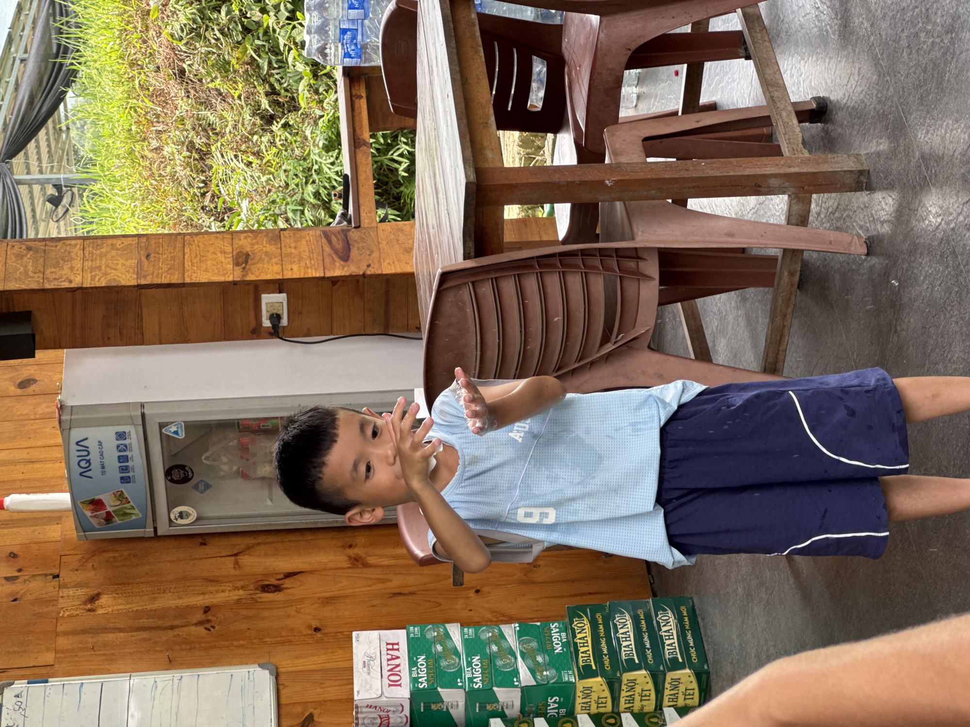 This image captures a bright, daytime scene in what appears to be a rustic indoor or semi-outdoor establishment in Du Già Commune, Vietnam. The main subject is a young boy, possibly between 4-7 years old, with short dark hair, wearing a light blue short-sleeved shirt with the number "6" and dark blue shorts. He is standing, facing slightly to his left, and is engaged in an activity with his hands, which are held up near his face, appearing to be wet or covered in a foamy substance, possibly from washing or playing with water.
The setting features wooden walls and a tiled floor. To the left of the boy is a white upright refrigerator with a glass door, displaying products inside. Beyond the wooden structure, through an opening, lush green foliage is visible, suggesting a natural outdoor environment. Several brown plastic chairs are stacked or arranged near a wooden table in the background. A partially visible adult's leg is seen in the lower right corner.
Notable details include:
- A power outlet on the wooden wall.
- Stacks of beer boxes on the floor, including brands like "HANOI" and "BIA SAIGON SPECIAL," with text like "CHÚC MỪNG NĂM MỚI" (Happy New Year) and "TẾT" (Lunar New Year), indicating a festive or retail context.
- The refrigerator has "AQUA" and "TỦ MÁT CAO CẤP" (Premium Cooler/Freezer) visible on its front.
- Water bottles on the table also bear the "AQUA" brand.
- A whiteboard in the lower left shows some handwritten notes, including a price "50k".
The overall impression is a casual, everyday scene in a local Vietnamese establishment, possibly a small shop or eatery.