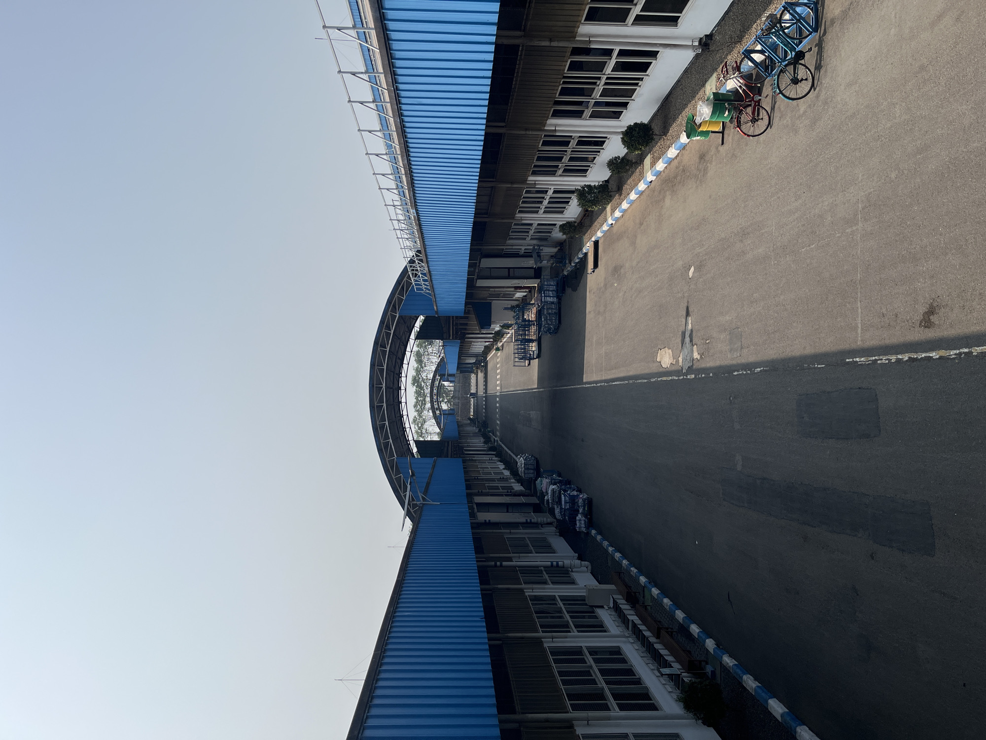 The image depicts a wide, paved roadway leading through a factory or industrial complex. The roadway is flanked on both sides by long, low-rise buildings with blue corrugated metal roofs and white walls. The buildings have numerous windows. A light-colored sky with a slight haze is visible above.
Along the right side of the roadway, near the foreground, are two bicycles parked in a blue rack, two green trash receptacles, and a red bicycle. Further down the road on the left, a line of trolleys loaded with goods is visible. Trees can be seen in the distance, beyond the industrial structures. The scene appears to be in Pelang, Indonesia, indicated by the context provided. The overall impression is one of an organized, operational industrial environment under a clear sky.
