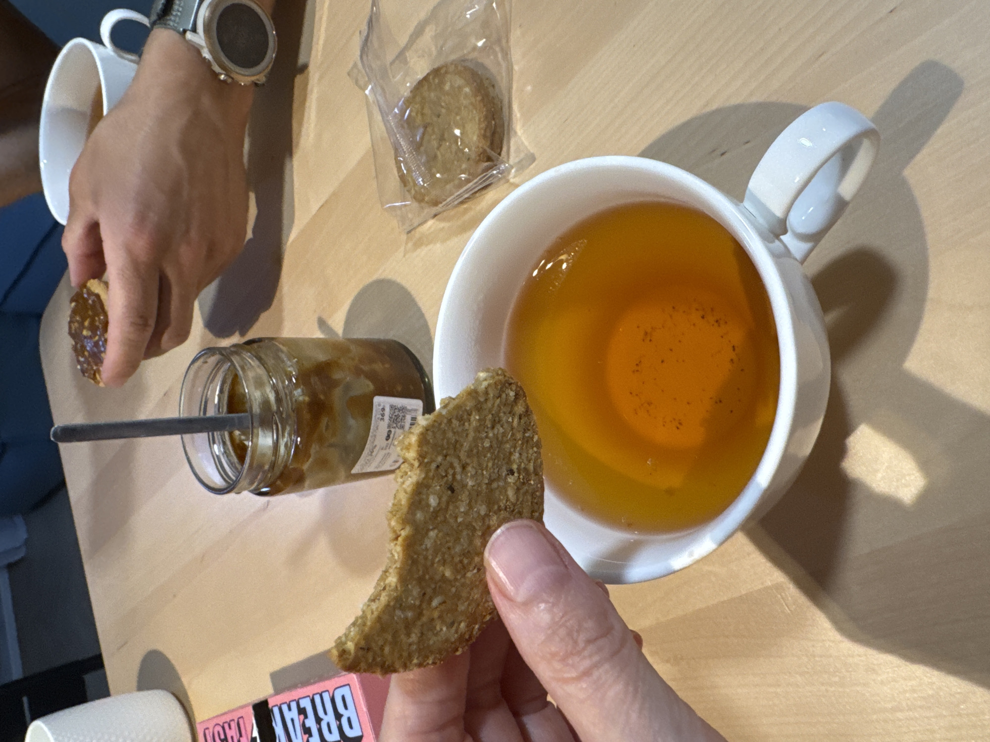 The image, taken in Izhevsk, Russia, captures an overhead view of two individuals enjoying a snack or light meal at a wooden table. Two hands are visible; one on the left, wearing a smartwatch with a light-colored bezel and dark strap, holds a round biscuit topped with a dark, possibly caramelized or fruit spread. The other hand, on the right, with a manicured thumbnail, holds a plain, light-brown biscuit, which appears to be oat-based or similar.
A white ceramic mug on the right contains an amber-colored liquid, likely tea, with small specks visible within. Another white mug is partially visible on the far left. A glass jar, possibly containing honey, jam, or a spread of a similar consistency, sits on the table with a spoon or stirrer inserted into it. A label on the jar shows the number "369". Another biscuit, individually wrapped in clear plastic, rests near the top center of the frame.
Strong shadows are cast across the light wooden table surface, indicating bright lighting, possibly from a window or overhead light source. Part of a pink box or packaging is visible in the lower-left foreground with the words "BREAK" and "FAST" printed on it, suggesting a breakfast item. The scene conveys a casual tea or snack break for two people.
