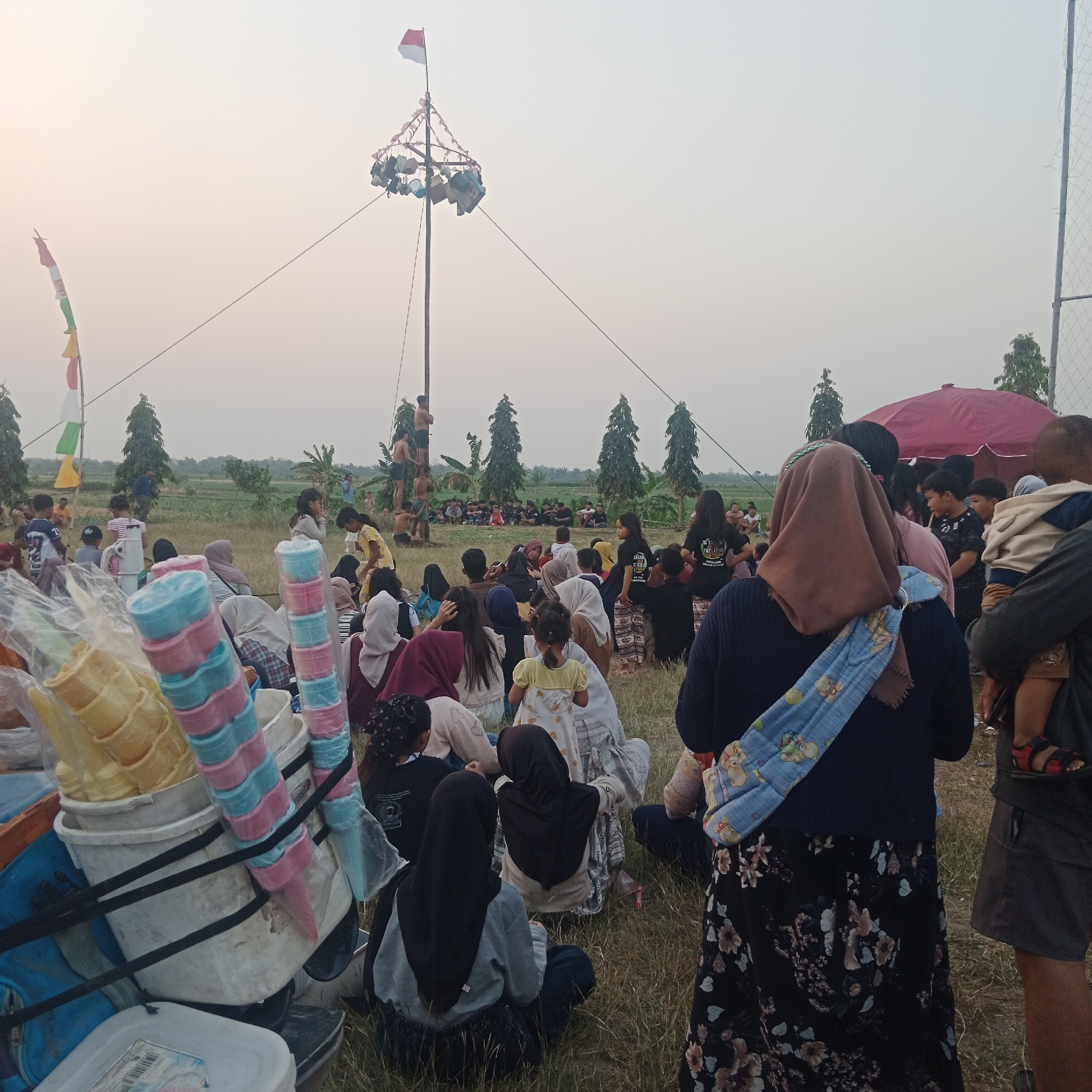 The image depicts an outdoor gathering in a grassy field, likely in Tegowanu Wetan, Indonesia, with a tall wooden pole at its center. The pole is adorned with what appears to be decorative elements and is topped with an Indonesian flag. Several individuals, predominantly young men, are positioned on and around the pole, suggesting some form of activity or competition. A crowd of people, including adults and children, are seated and standing in the foreground and midground, observing the scene. Some are facing the pole, while others are looking in different directions. A stall or cart with stacks of colorful ice cream cones is visible in the lower left of the frame, indicating a potential vendor presence. The environment appears to be a rural or semi-rural area, with sparse trees and vegetation in the background, under a hazy sky. The overall atmosphere suggests a community event or festival.