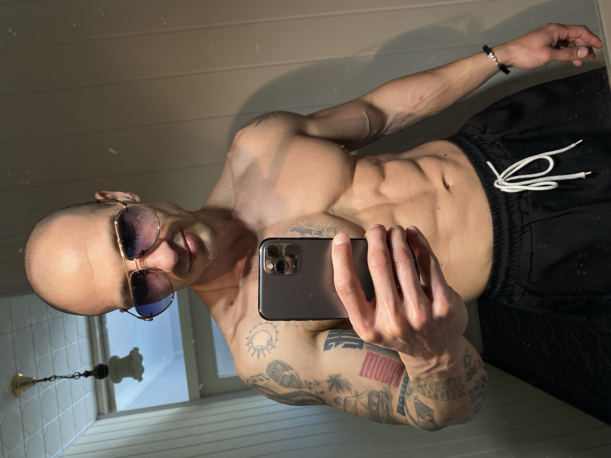A shirtless man with a shaved head and sunglasses is taking a selfie with a smartphone. He has multiple tattoos on his arms and torso. He is wearing black shorts. The setting appears to be a bathroom with tiled walls and a window reflecting daylight. A light fixture is visible in the reflection. There is no discernible evidence to definitively determine the location as Pleven, Bulgaria.