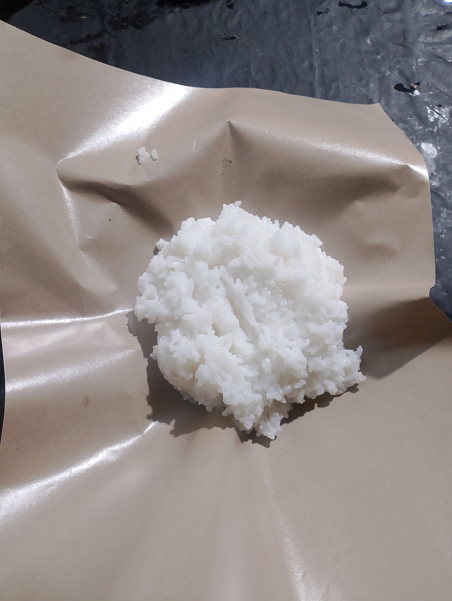 A pile of cooked white rice sits on a sheet of brown paper, which is placed on a dark, textured surface. The rice is slightly moist and clumps together. The brown paper is crumpled and reflects light in some areas, suggesting it is a thin, possibly waxed or coated paper. The dark surface underneath appears to be a tabletop or ground, possibly outdoors given the lighting. There are no people or other objects visible in the immediate vicinity. The setting is likely a casual outdoor or indoor environment where food is being served or prepared in a simple manner. The image is taken from a slightly elevated angle looking down. The overall scene depicts a simple serving of rice, common in Indonesian cuisine. The specific location in Kedungsari, Indonesia, suggests a local culinary context.