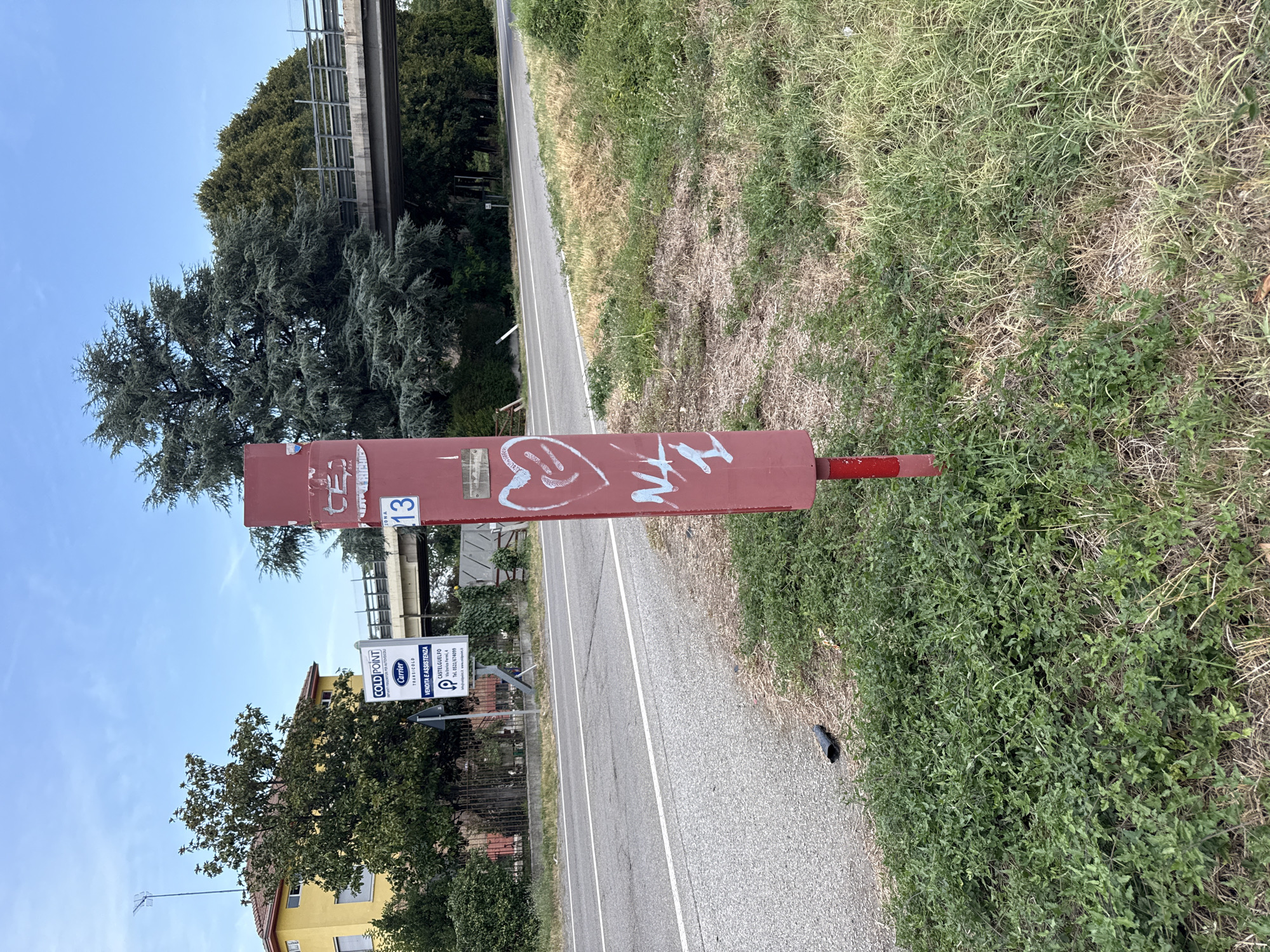 The image shows a red metal post with graffiti in the foreground, positioned on the side of a road in Fontevivo, Italy. The post has a white drawing of a heart with squiggly lines inside, and what appears to be the letters "NL 1" also in white. Further up the post, there is a small white sticker with some text and a blue emblem. To the left of the post, a road is visible with white lane markings. In the background, there is a yellow building with a tiled roof, a sign for "COLD POINT" with the Carrier logo, a green tree, and a bridge or overpass. The environment is outdoors, with grass and dry vegetation surrounding the post and the road. The sky is blue with some white clouds. A small black object, possibly a piece of trash, is on the ground near the road.