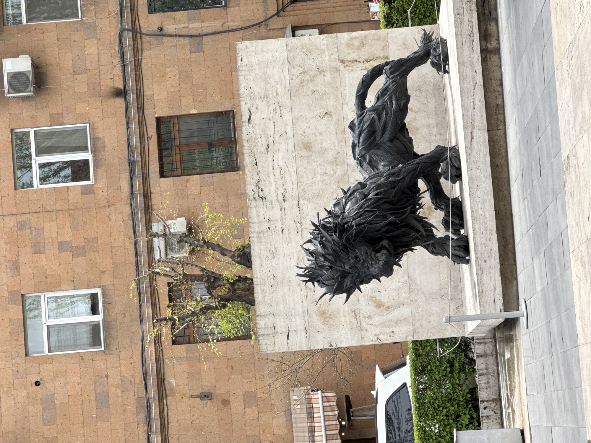 The image shows an exterior view in Yerevan, Armenia, featuring architectural elements and a sculpture. A light brown building with windows and an air conditioning unit is visible on the left. Adjacent to it, a travertine-clad building features a sculpture depicting a dark, mythical creature or lion-like figure in a dynamic pose. The sculpture is placed against a travertine wall, and part of a tree with budding leaves is visible in the foreground. The background includes urban elements such as a street, a white vehicle, and additional buildings. The image is taken from an elevated perspective.