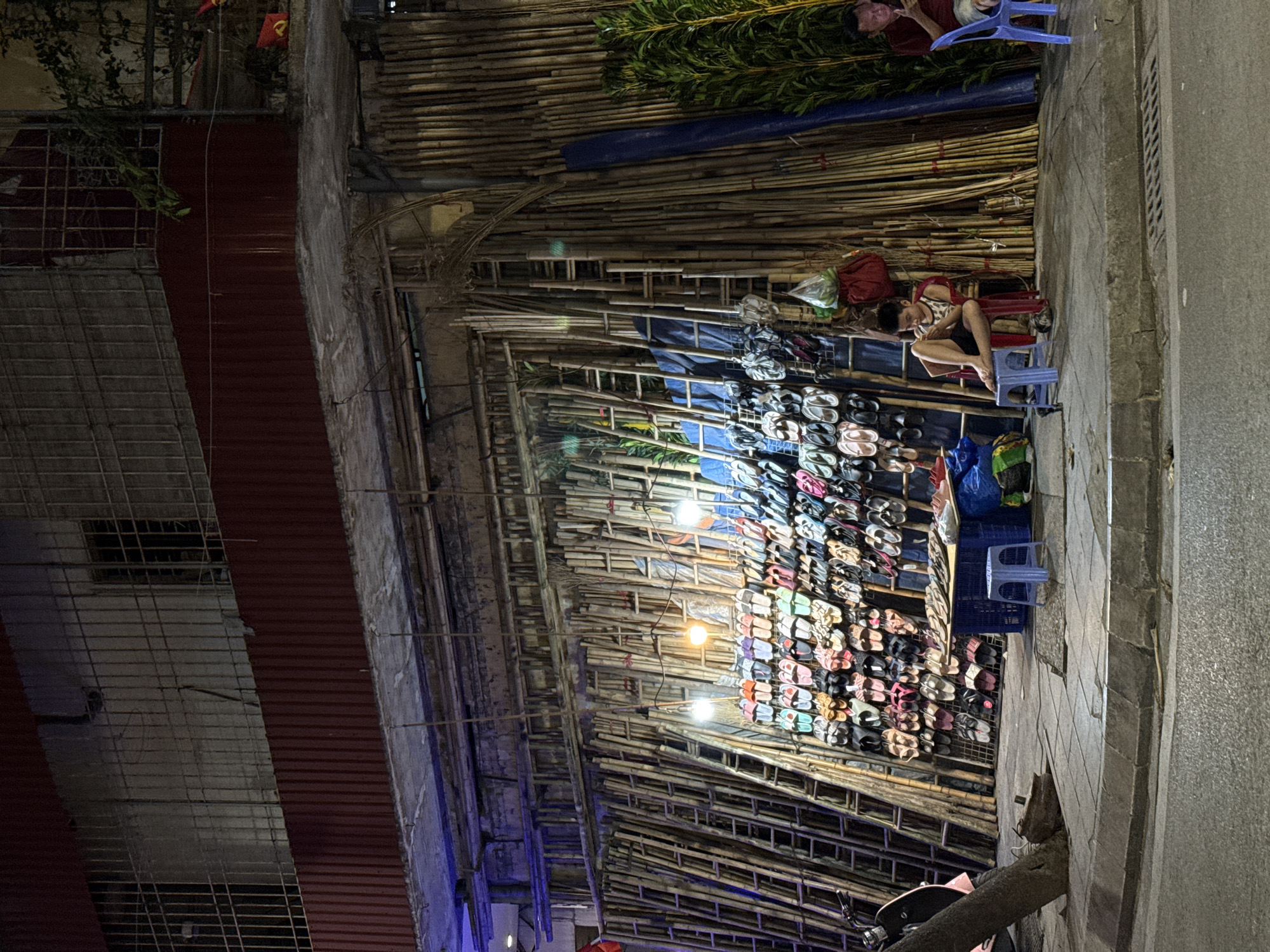 The image, oriented correctly, depicts a lively outdoor market stall at night, likely in Hà Nội, Vietnam. The scene is illuminated by bare light bulbs strung across a unique, predominantly bamboo structure that forms the backdrop and canopy of the stall. This structure consists of numerous bamboo poles stacked horizontally, creating a textured wall, with some sections covered by blue tarpaulins and adorned with long green foliage.
The main subject of the stall is a display of numerous pairs of flip-flops and sandals, arranged on metal racks, showcasing a variety of colors and designs.
Two individuals are present, both seated on small blue plastic stools. One person, positioned to the right of the center, wears a light-colored, patterned short-sleeved top and dark shorts, looking down as if resting or engaged with a personal device. Another person is partially visible in the upper right quadrant, seated on a similar stool behind a section of green foliage, wearing a red shirt.
The ground consists of paved stone tiles, and a metal drainage grate is visible near the street edge. On the left side of the image, urban architecture is visible, including a building with a red corrugated roof and barred windows. A red flag with a yellow star, identifying it as the national flag of Vietnam, can be seen in the upper left background, partially obscured by foliage. A portion of a motorcycle or scooter, including a wheel and handlebars, is visible in the bottom right corner, parked near the curb.
The time of day is clearly night, indicated by the dark surroundings and the dependence on artificial light sources. No legible text is visible in the image. The overall impression is that of a typical evening street market in Southeast Asia.