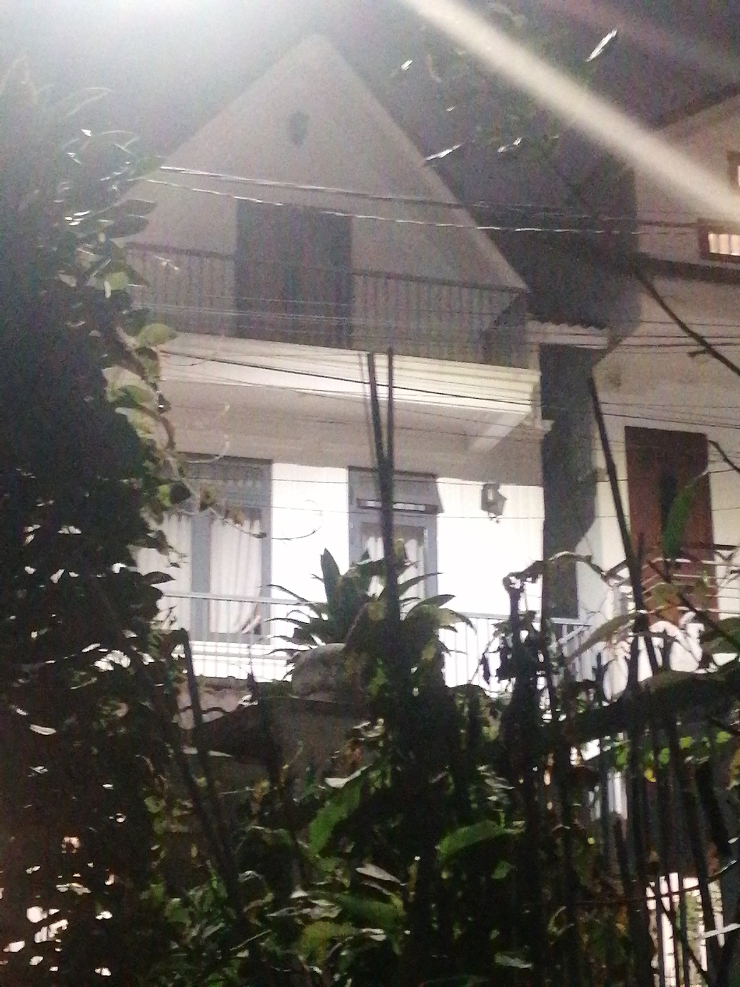 The image depicts a facade of a multi-story building, likely residential, at night. The building features balconies, windows with white curtains, and a prominent gable roof. Dark foliage, possibly trees or bushes, obscures the lower portion of the building and parts of the upper levels, creating a sense of depth and a slightly overgrown environment. Overhead wires are visible against the bright sky, indicating an urban or suburban setting. The lighting suggests artificial illumination, with bright white light creating lens flares and washing out some details. The location is identified as Phường Xuân Hương - Đà Lạt, Vietnam.