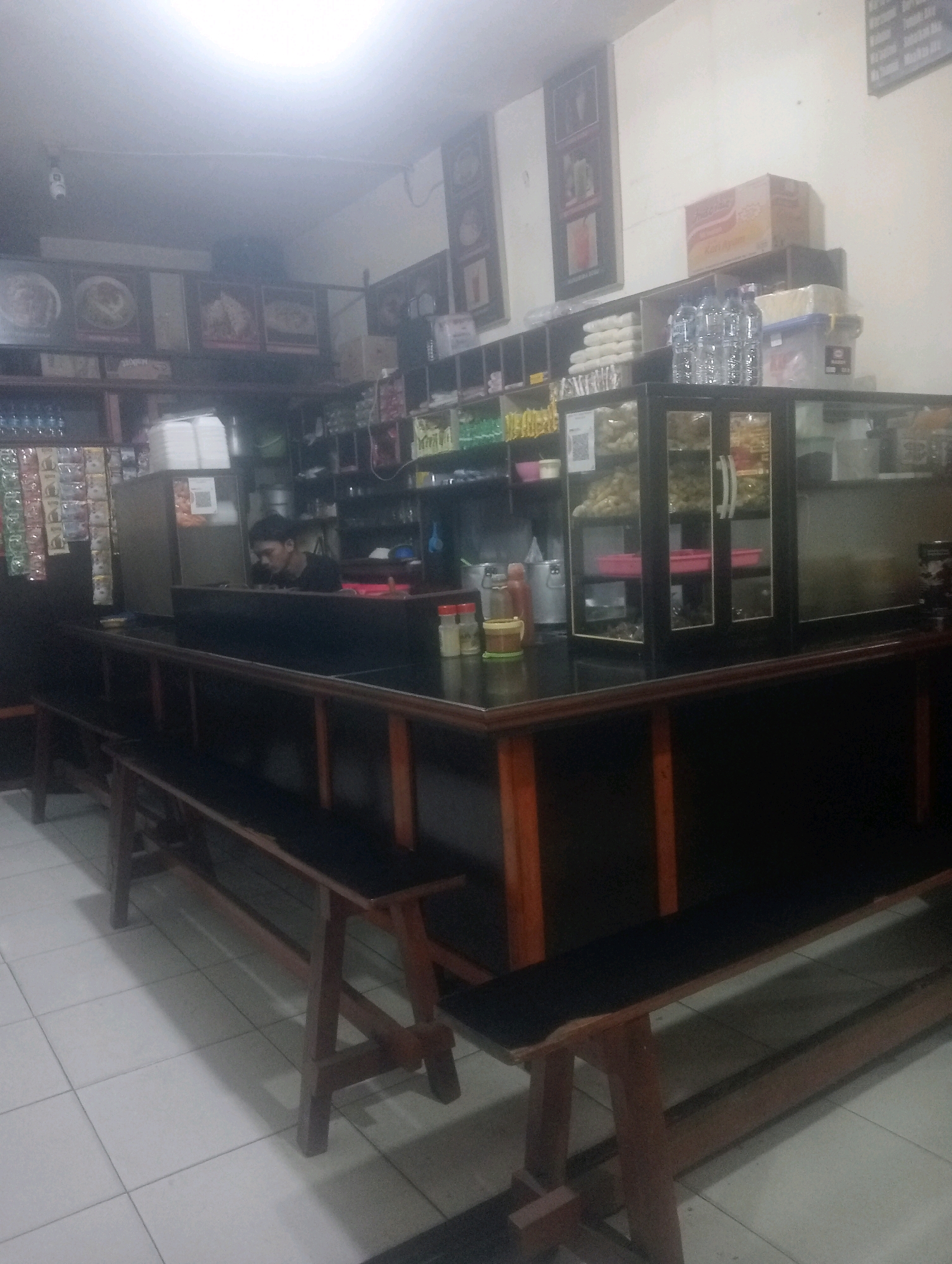 The image shows the interior of a small, dimly lit food stall or cafe, likely in South Jakarta, Indonesia. A person, appearing to be a male employee wearing a black shirt, is visible behind a counter, looking down. The counter is dark and appears to be made of wood, with a glass display case on top filled with various food items. To the left of the counter, a shelf unit holds packaged snacks and drinks, with a stack of white containers on top. Further to the left, a display of bottled water is visible. In the foreground, wooden benches with dark seating are arranged on a tiled floor. The walls are adorned with framed pictures and shelves stocked with items. The overall impression is of a functional, no-frills establishment.