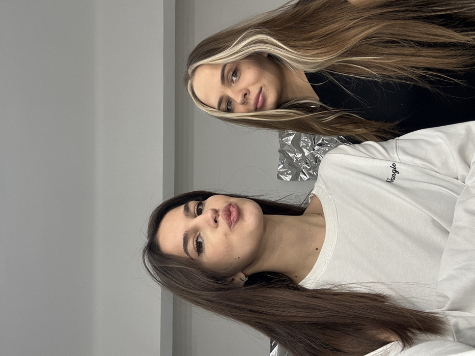 Two women are posing for a photo. The woman in the foreground has dark brown hair, plump lips, and is wearing a white long-sleeved shirt. The other woman behind her has blonde hair with lighter blonde highlights and is wearing a black shirt. Both women are facing forward. Aluminum foil is visible between them. The background is a solid, light gray color. The image is slightly tilted.