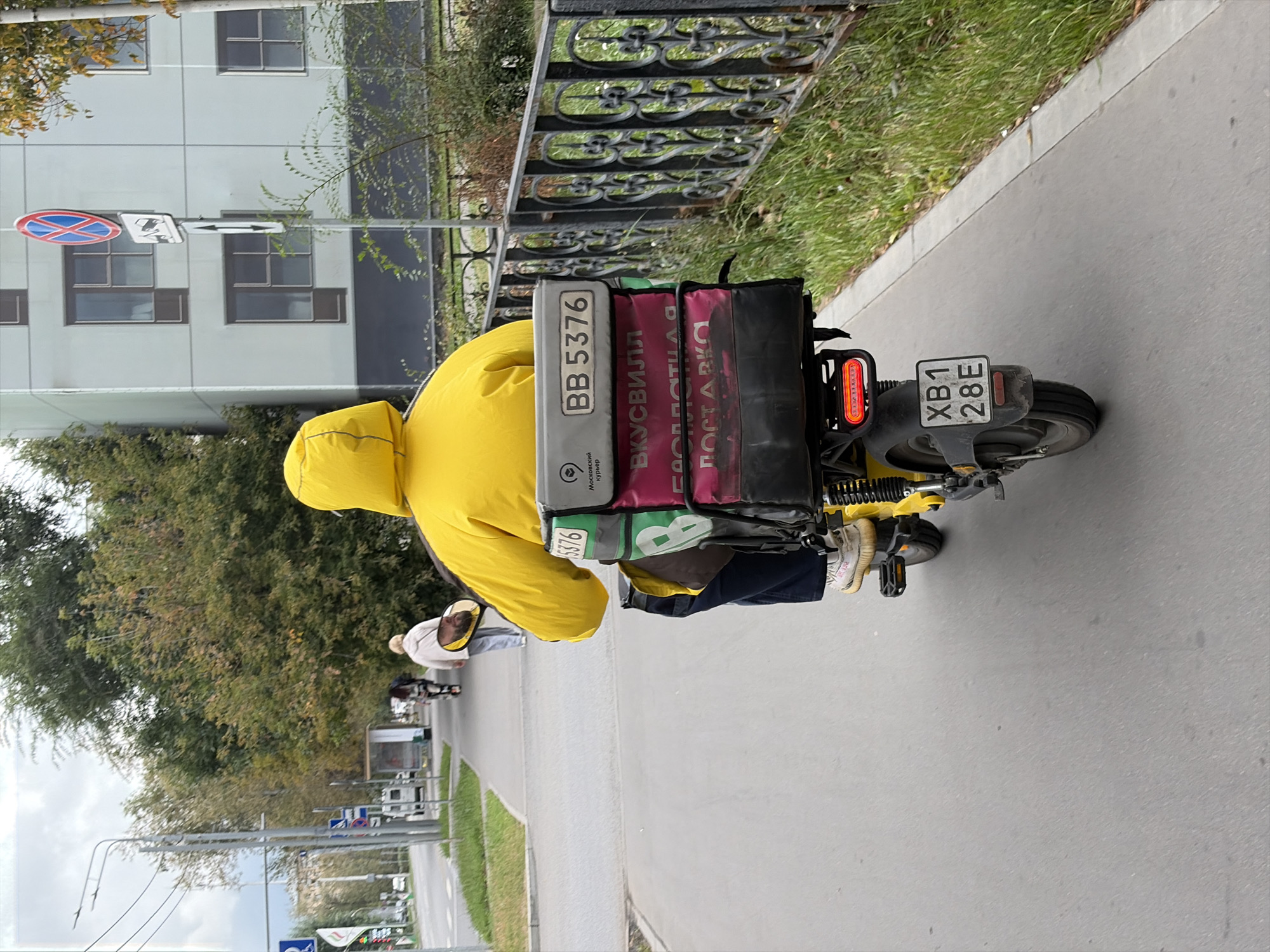 A person wearing a bright yellow hooded jacket is riding a yellow electric scooter down a paved sidewalk. The scooter has a large black delivery box attached to the back, which is adorned with a white license plate reading "BB 5376" and red lettering. The box also has a logo that appears to be a stylized "B" or similar symbol. Further down the box, red lettering is visible that reads "ВКУСВИЛЛ" and "ДОСТАВКА," suggesting it is a delivery service for the VkusVill brand, a Russian supermarket chain. The scooter has a license plate at the rear reading "XB1 28E." The sidewalk is bordered by a black metal fence and grassy areas. In the background, a modern building with multiple windows is visible, along with trees and a traffic sign. The sky is overcast. The scene appears to be a street in Moscow, Russia, based on the Cyrillic text and the general urban landscape.