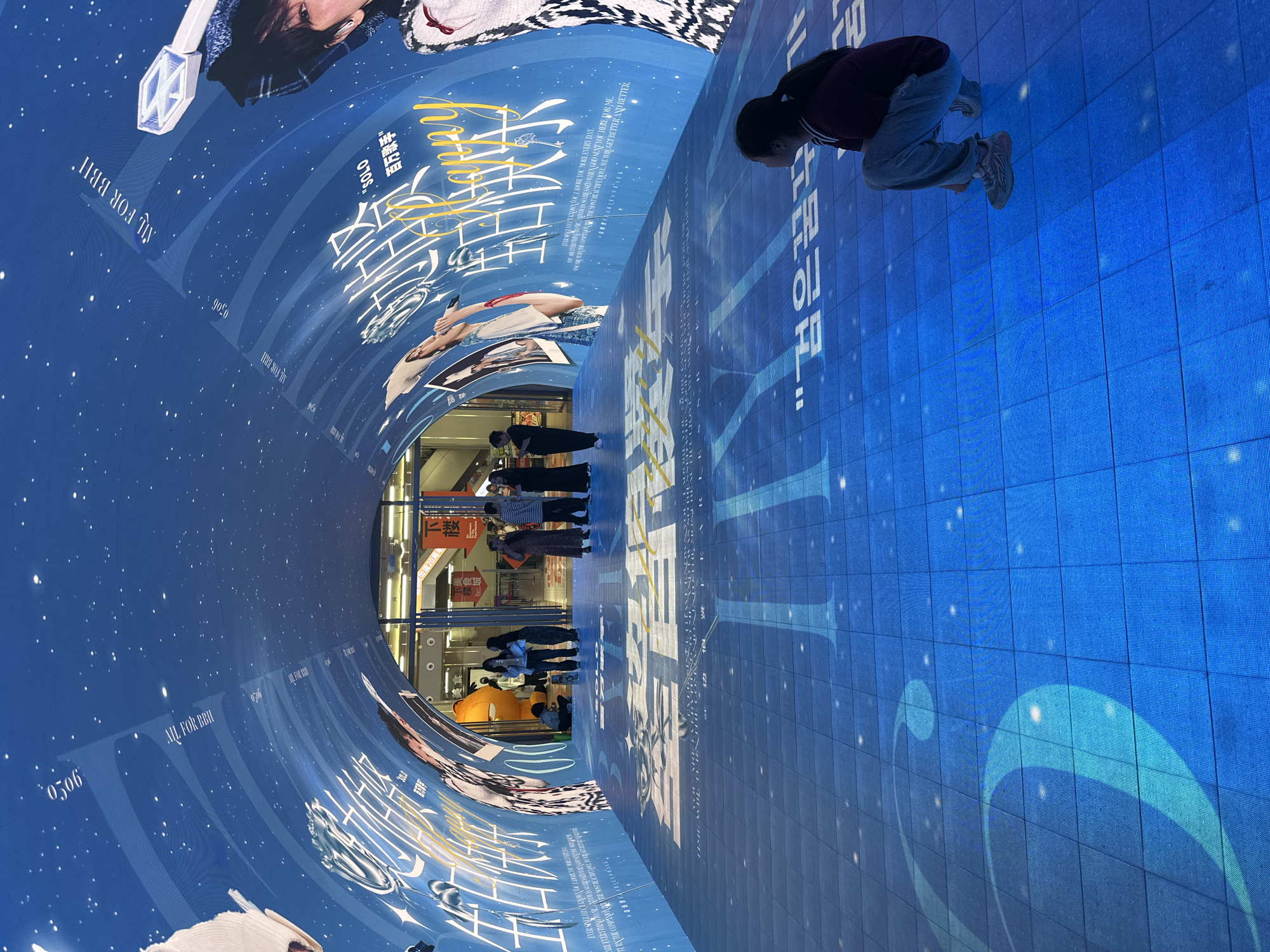 The image shows an indoor space, possibly a commercial area in Yuzhong District, China, based on the Chinese characters on the posters. The space has curved walls and flooring with a blue hue, seemingly lit by a large digital screen showing a design resembling a starry sky. The screen displays images of a person (presumably a celebrity) and text in Chinese and English, including phrases like "ALL FOR BBH" and "SOLO." Several people are standing in the distance, observing the display. A woman is kneeling closer to the foreground, appearing to interact with the floor screen. There is signage visible in the background, including characters for "food street."