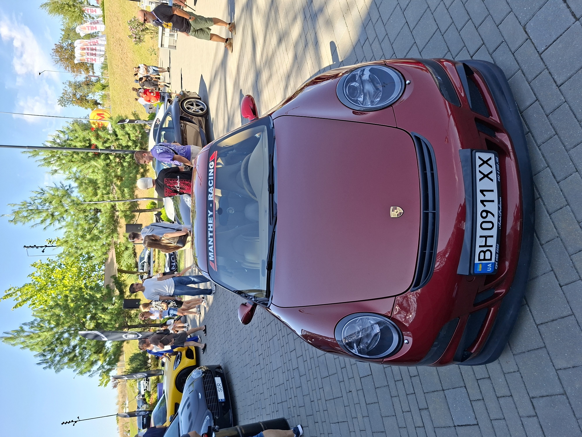 In Stăuceni, Moldova, on a sunny day, a red Porsche 911 is parked on a cobblestone surface, its front facing the camera. The car has a black bumper and a license plate that reads "BH 0911 XX". A banner with "MANTHEY-RACING" is visible on the windshield. Several people are gathered around other cars, including a yellow sports car and a black sports car. The background features green trees, a hill with banners, and a clear blue sky. Some individuals are walking and observing the vehicles.
