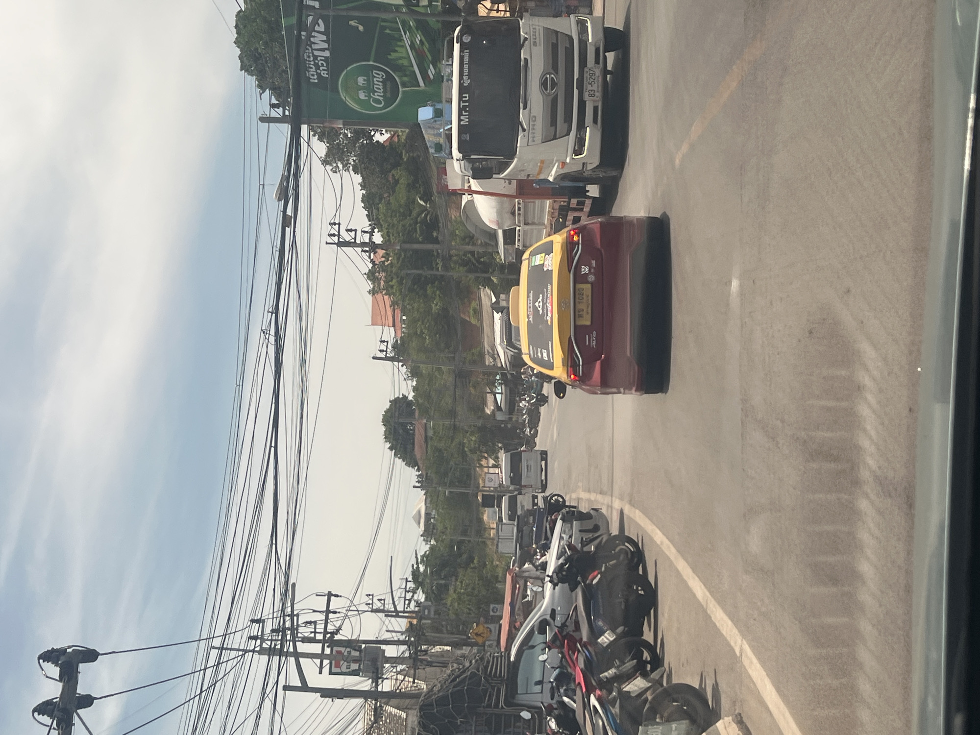 The image captures a street scene in the Ko Samui District of Thailand. The perspective is from inside a vehicle looking forward. On the right side of the road, a white Hino truck with a crane is visible, bearing the name "Mr.Tu" on its windshield. Next to it, a red and yellow taxi is driving in the same direction. The road is lined with parked motorcycles on the left. Overhead, a complex network of electrical wires stretches across the sky, supported by utility poles. In the background, buildings and lush green trees can be seen. A sign for "Chang" beer is prominently displayed on the right side of the road. The sky is partly cloudy.