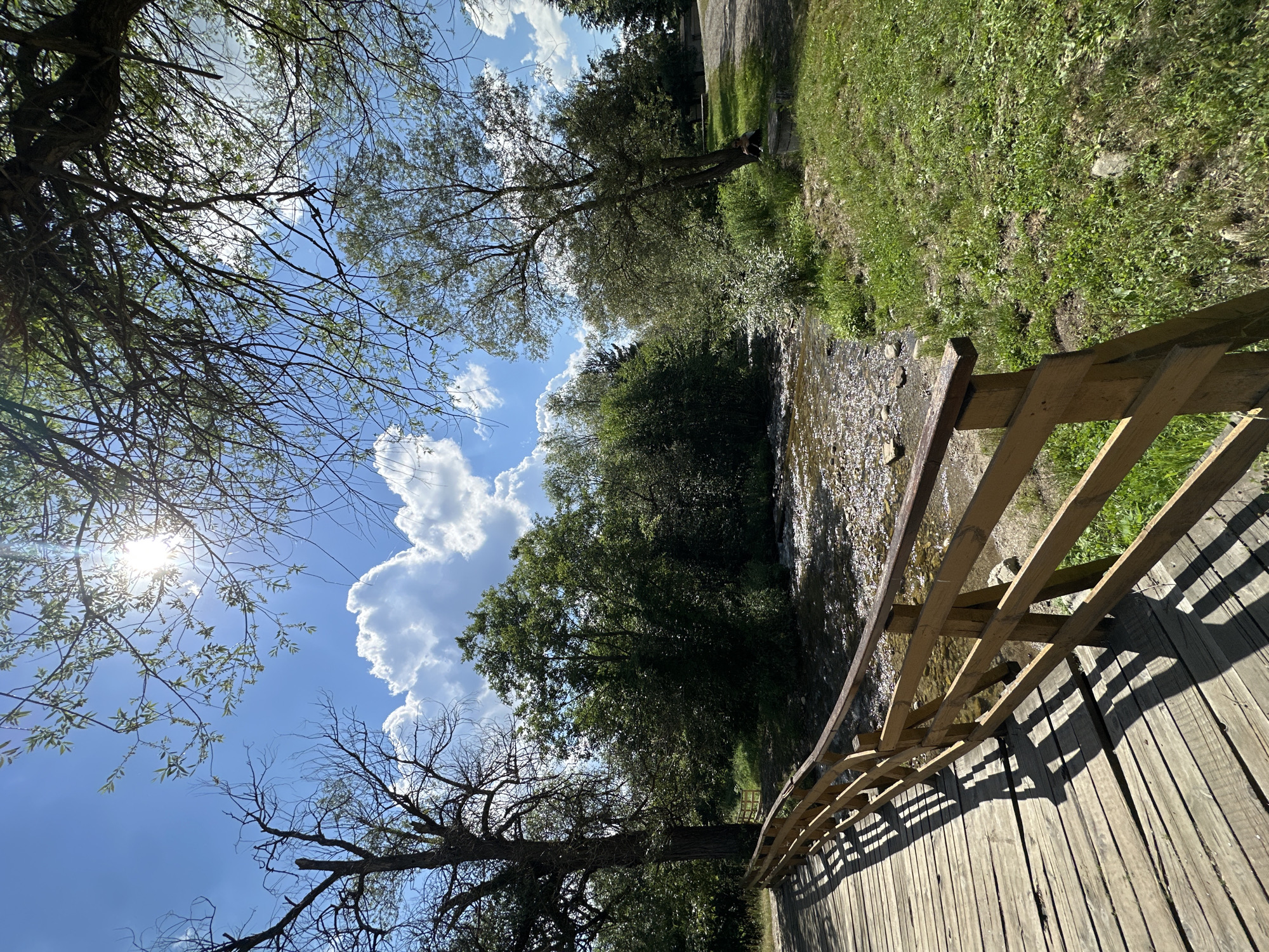 The image depicts a natural scene with a wooden bridge in the foreground. The bridge is made of weathered planks and has a wooden railing. Beyond the bridge, a shallow, rocky stream flows, with lush green trees and vegetation lining its banks. The sky is blue with scattered white clouds, and the sun is visible, casting a bright light and shadows. The overall setting appears to be a park or natural area. While the image captures a serene outdoor environment, there are no specific landmarks or clues that would definitively place it in the city of Samokov, Bulgaria. The scene is generic enough to be found in many similar natural locations.