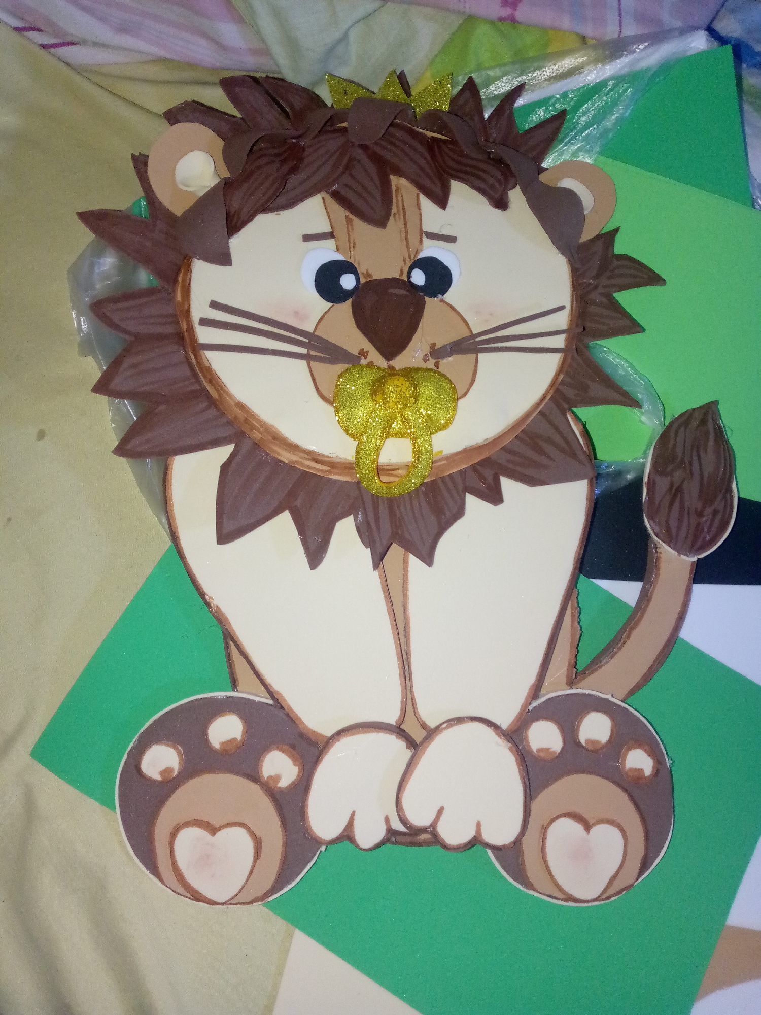 A cartoon lion with a yellow glitter pacifier is lying on a bed. The lion is made of brown and cream-colored foam. It has a dark brown mane and tail, cream-colored body, and brown paws with heart-shaped pads. The background includes pieces of green and white foam. There is no evidence in the image to confirm the location is Juan Griego, Venezuela.